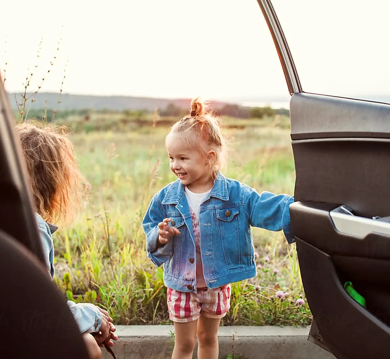 A young girl in a denim jacket stands by an open car door, smiling at another child—dreaming big with help from credit union loans—while the sun sets over the field.