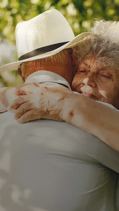 An elderly woman with curly gray hair hugs an elderly man wearing a white hat, both showing affection outdoors in sunlight.