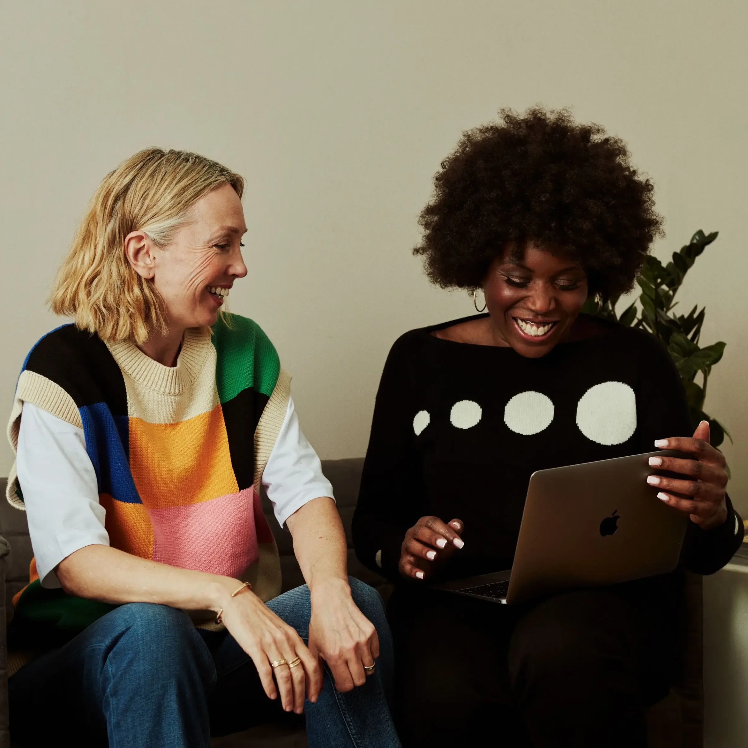 Two women sit on a couch, smiling at a laptop together—perhaps exploring the benefits of opening a money market account.