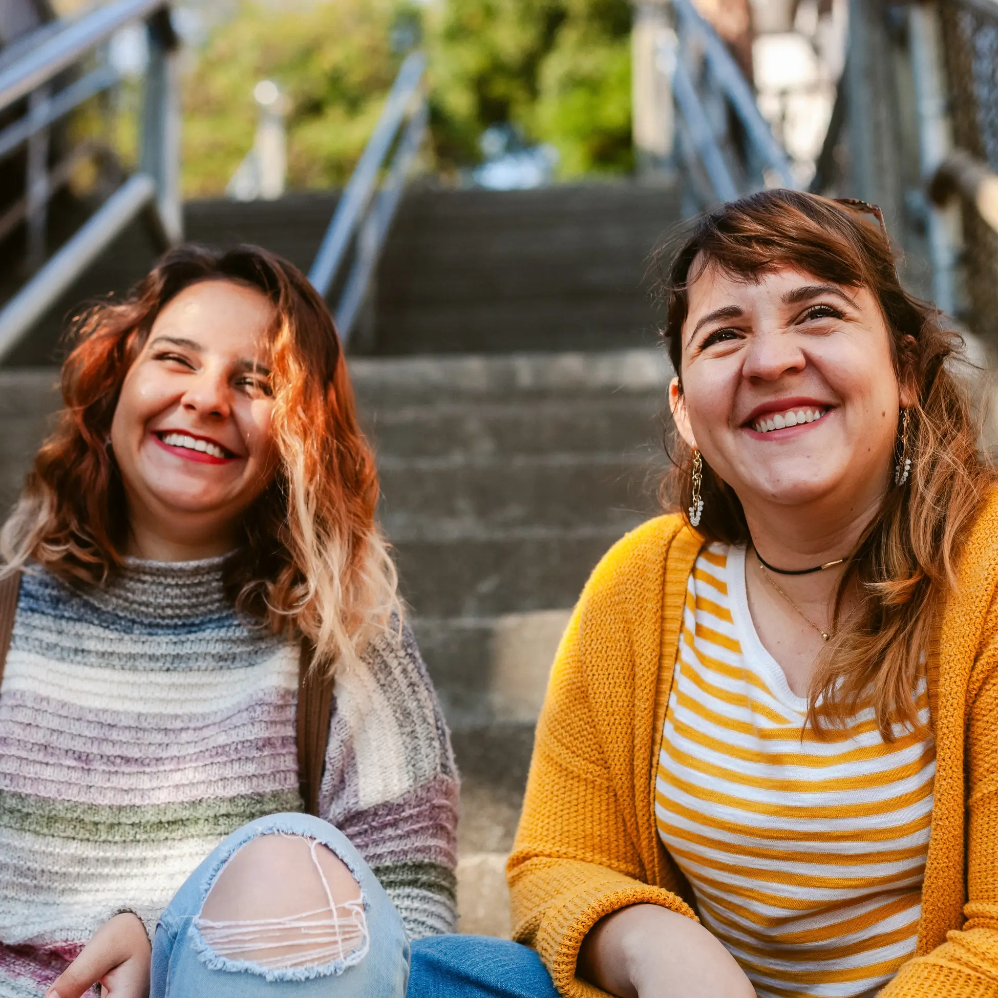Two women sit on outdoor steps, laughing together. Their joy shines like opening a credit union savings account—bringing warmth, comfort, and a brighter future under the trees in the background.