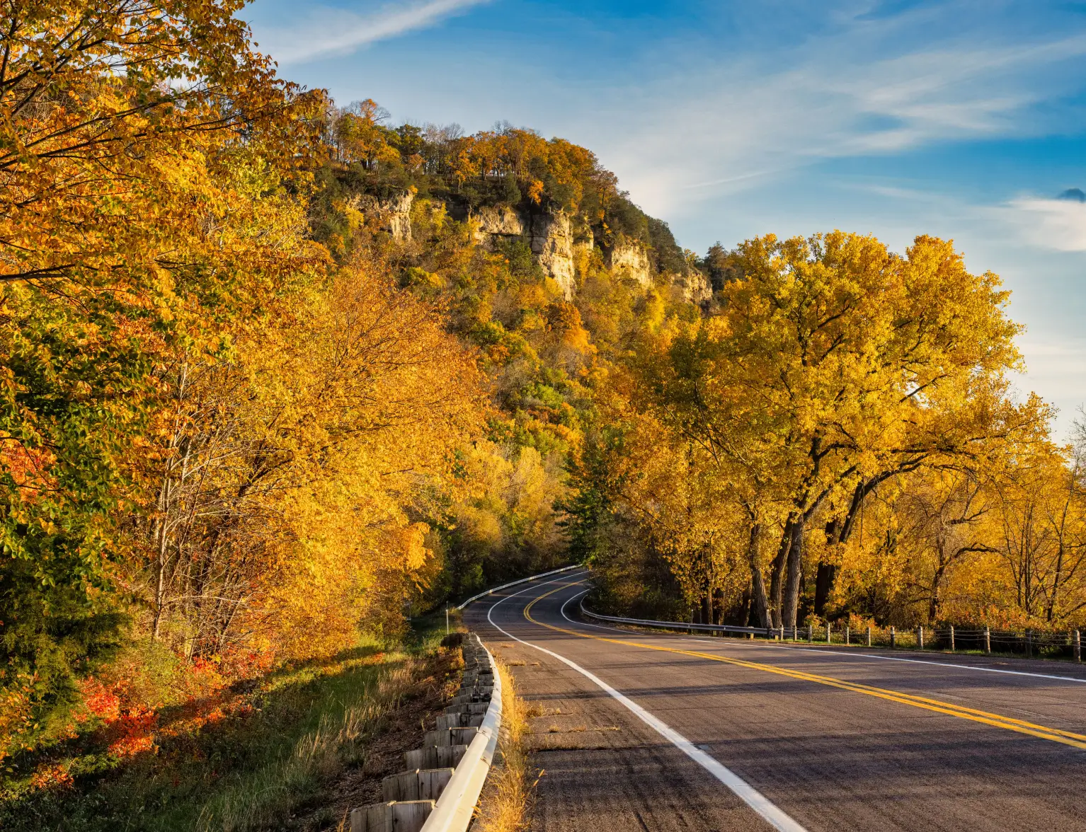 A winding road curves through a forest of golden autumn trees with rocky cliffs in the background under a blue sky.