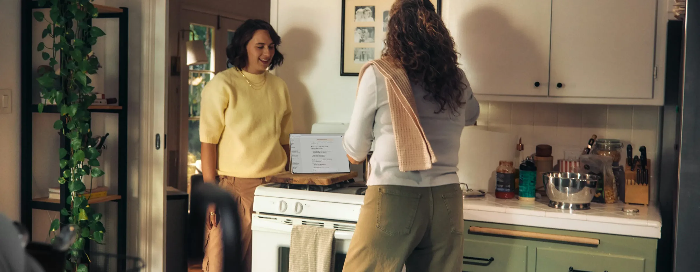 Two women cook together in a bright kitchen, one checking how to join a credit union on her laptop near the stove, surrounded by plants and utensils.