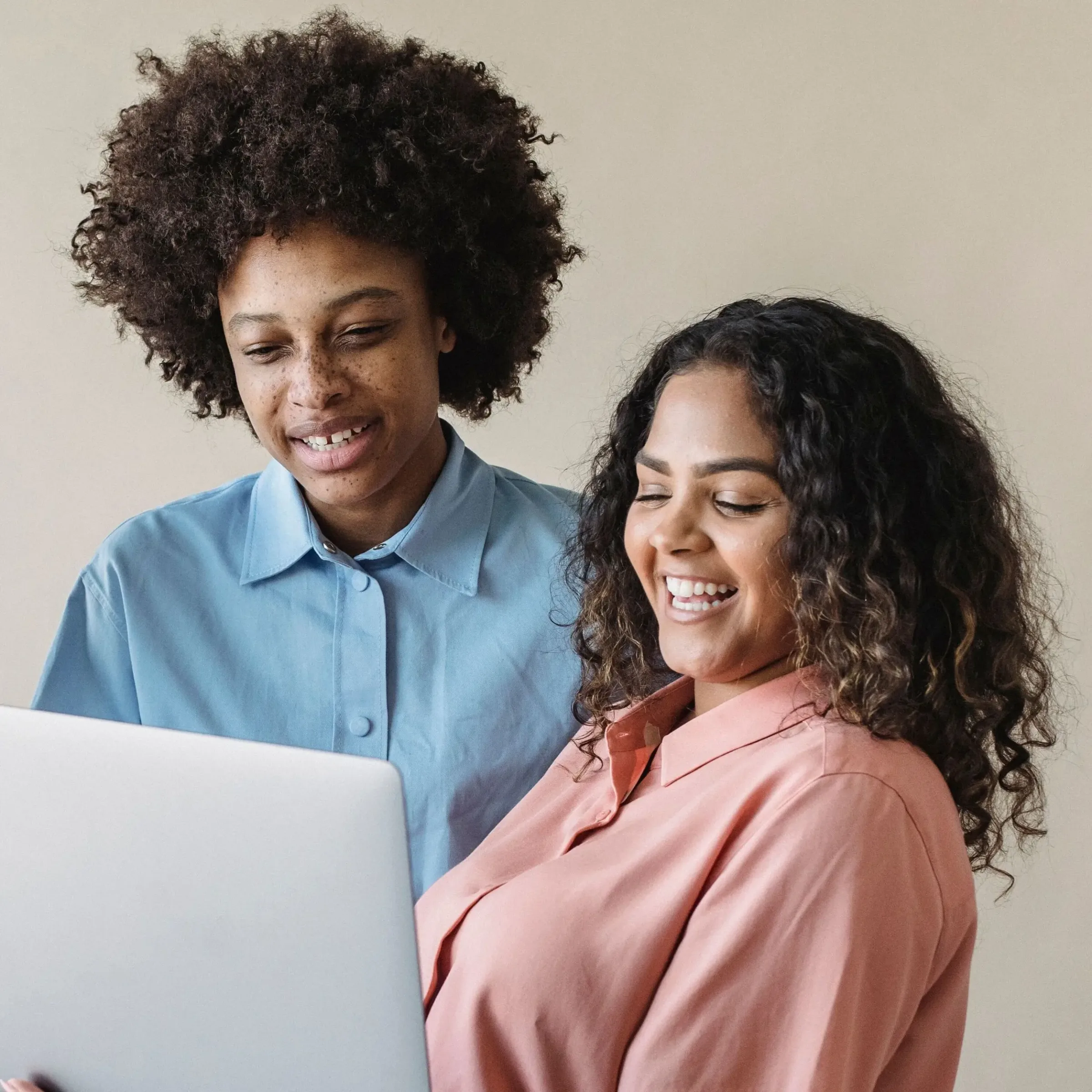 Two people smiling while looking at a laptop together, exploring credit union credit cards, both in button-up shirts against a plain beige background.