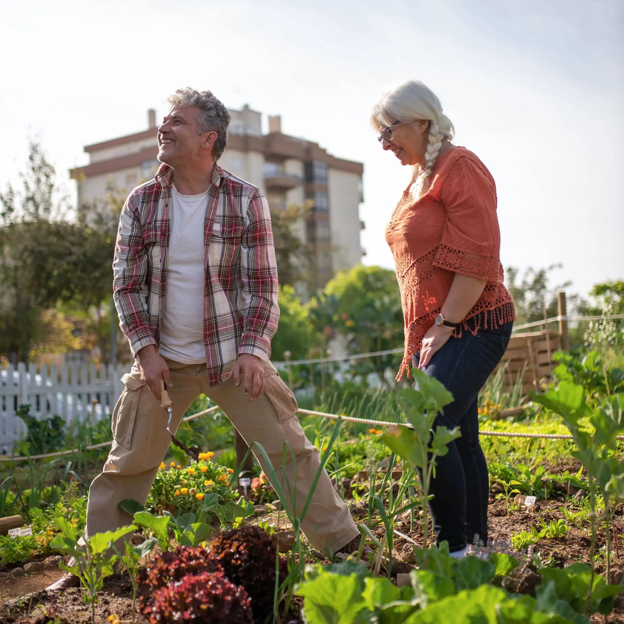 An older man and woman garden together, smiling and tending to plants in a sunny vegetable patch, chatting about their money market account as they nurture both their crops and savings.