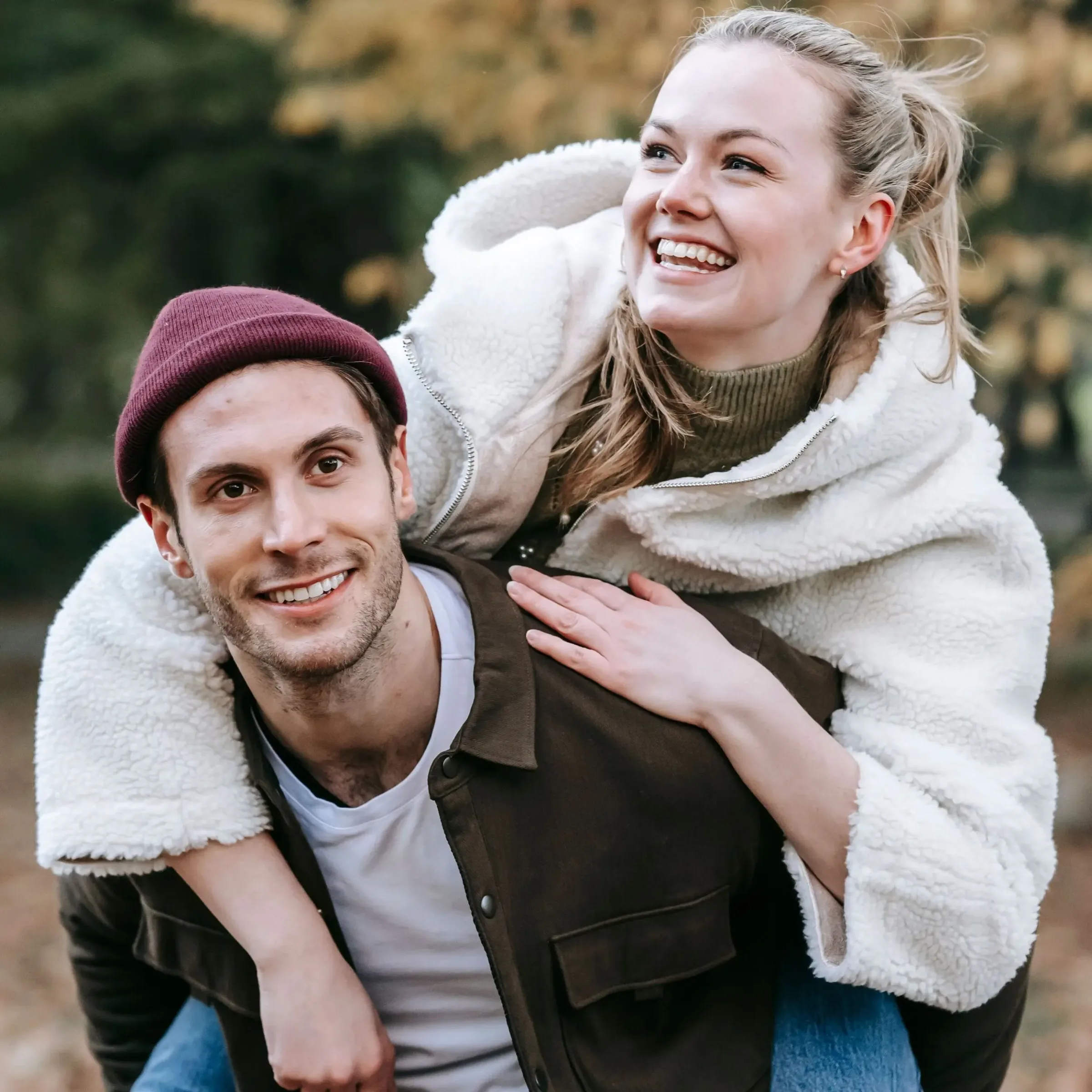 A smiling man in a brown jacket carries a woman on his back outdoors, both happy—like finding the perfect credit union checking account together.