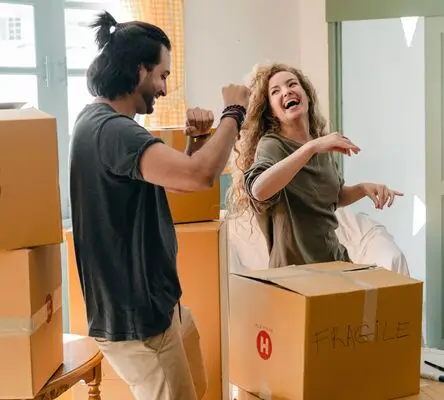 Two people smile and dance together among cardboard boxes in a bright room, suggesting they are moving into a new home.