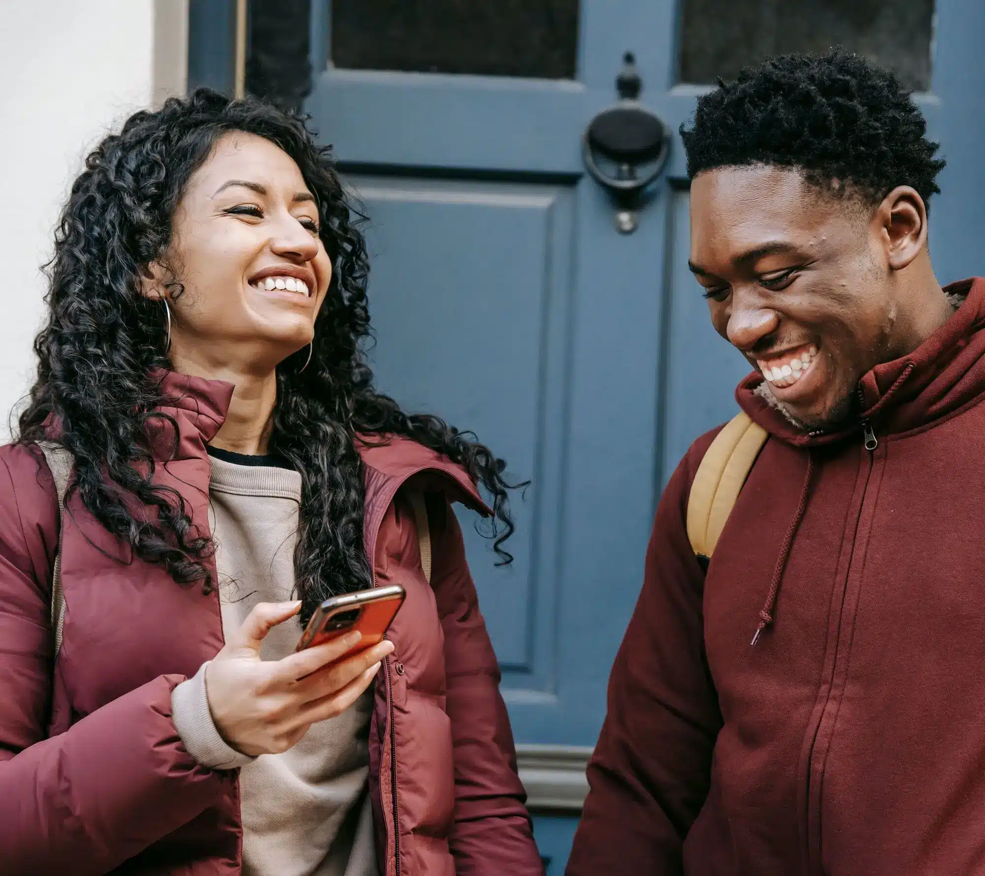 Two people in maroon jackets stand outside a blue door, laughing as they check a smartphone—perhaps to see if they’ve just won the saver’s sweepstakes together.