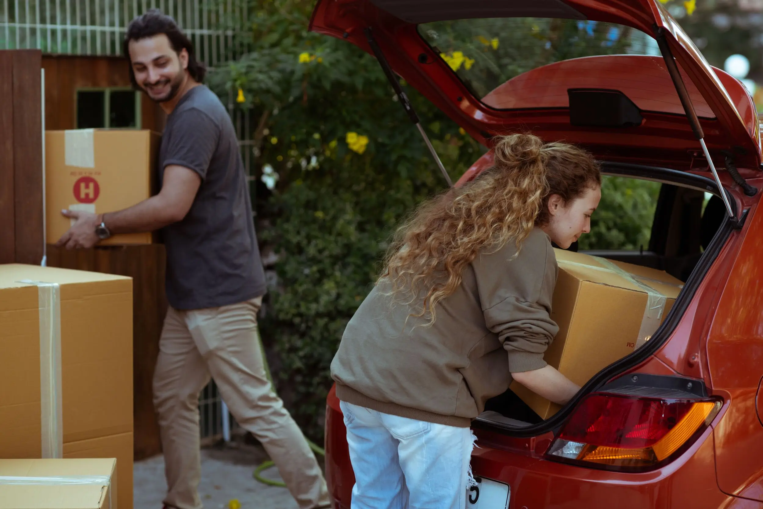 Two people load cardboard boxes into the trunk of a red car, perhaps moving to take advantage of low auto loan rates. Greenery and a fence form the background.