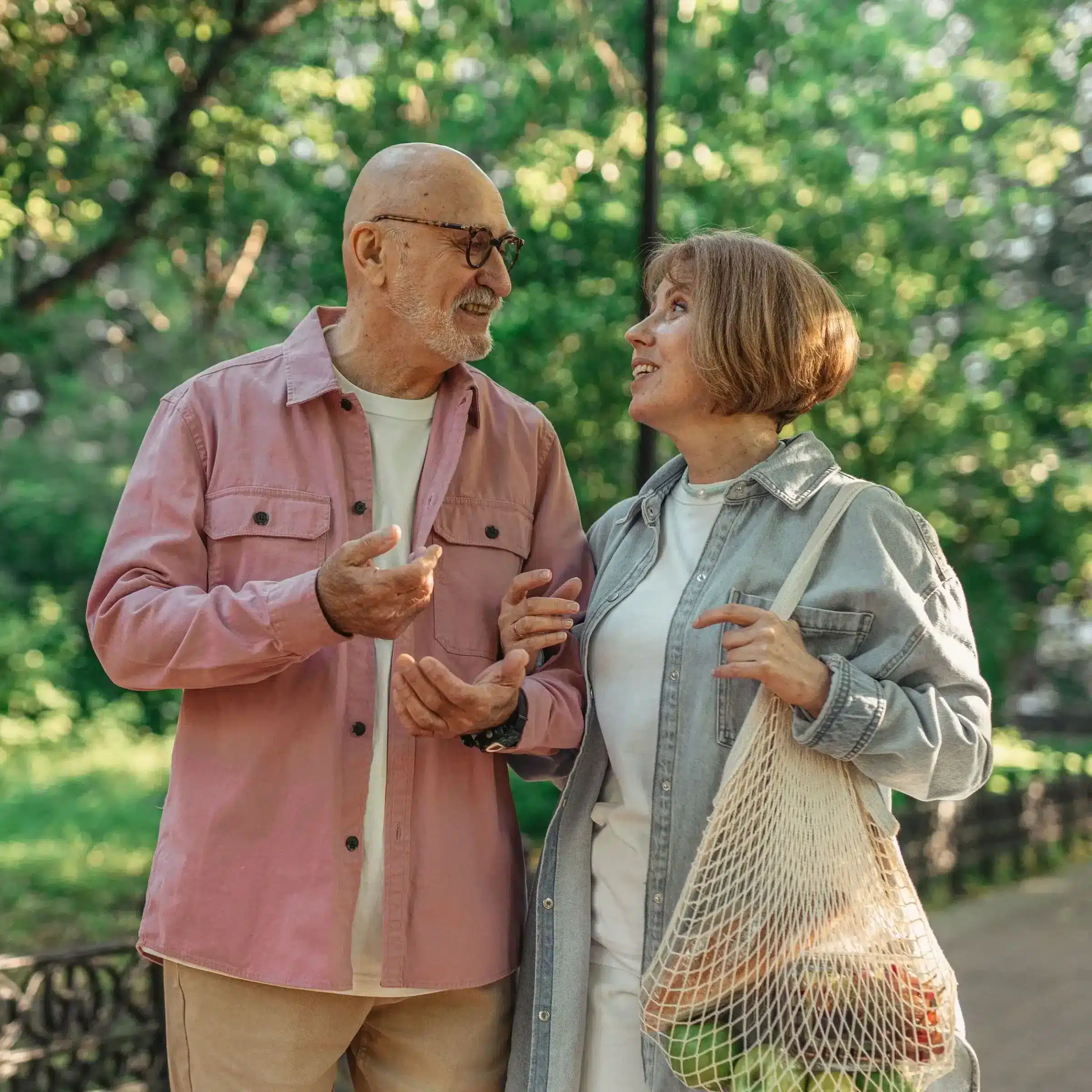 An older man and woman smile at each other while walking outdoors in Green Bay. The woman carries a mesh bag of groceries as sunlight filters through green trees in the background.