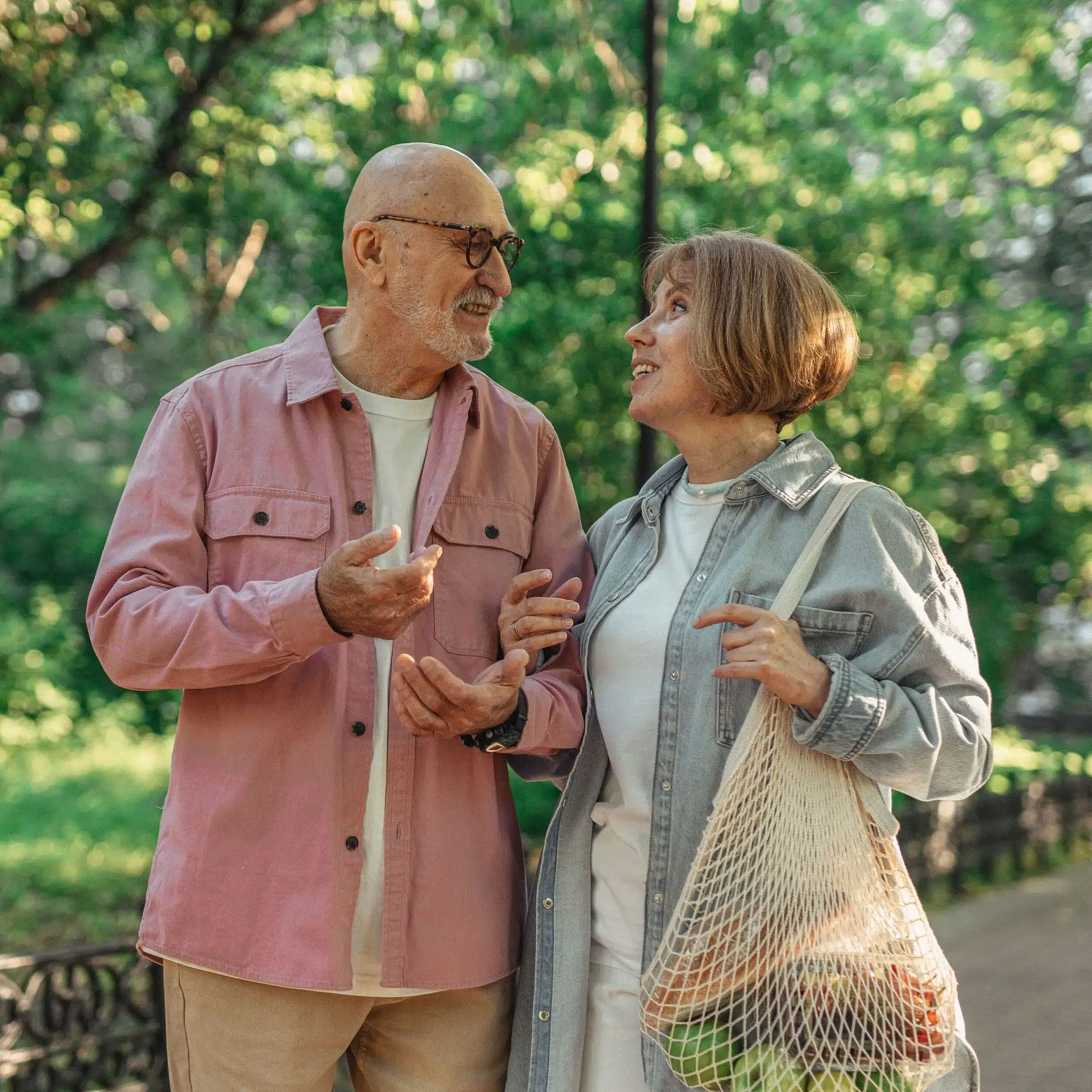 An older man and woman smile at each other while walking outdoors in Green Bay. The woman carries a mesh bag of groceries as sunlight filters through green trees in the background.