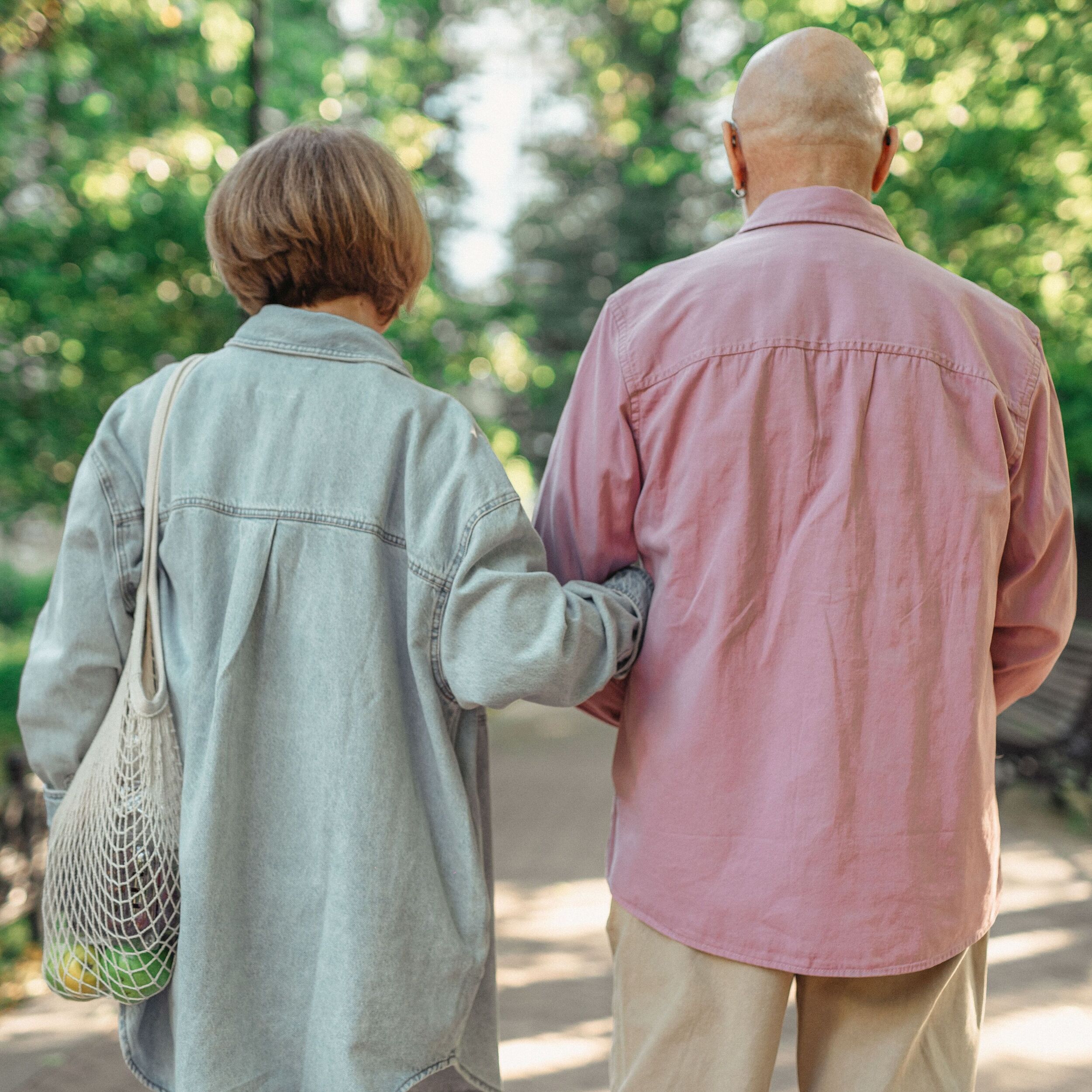 Two older adults stroll outdoors, one in denim with a mesh bag—perhaps after discussing their IRA account—while trees and greenery frame their peaceful walk.