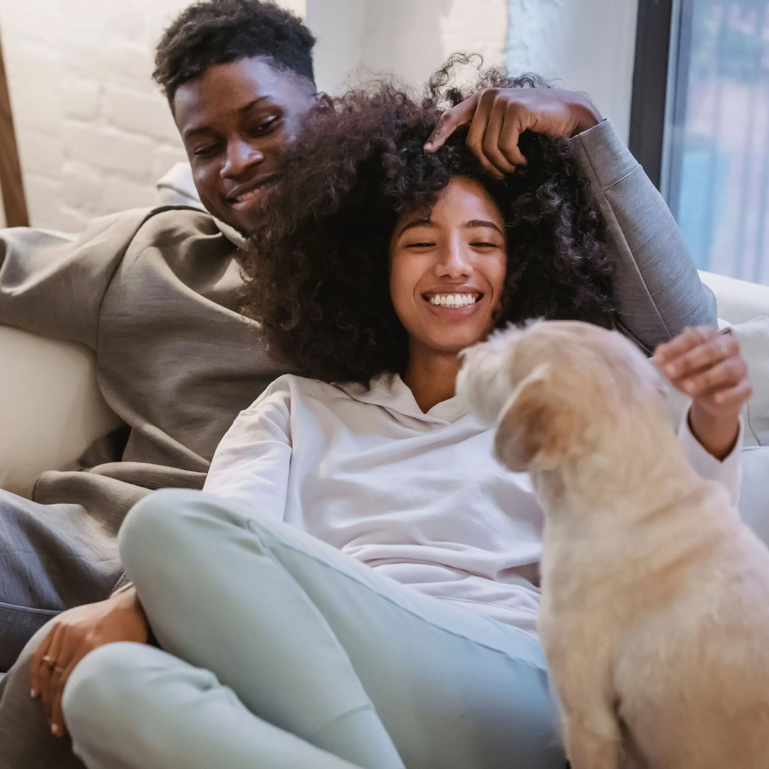 A smiling couple sits on a couch, playfully relaxing with their dog—dreaming about homeownership with an FHA loan.