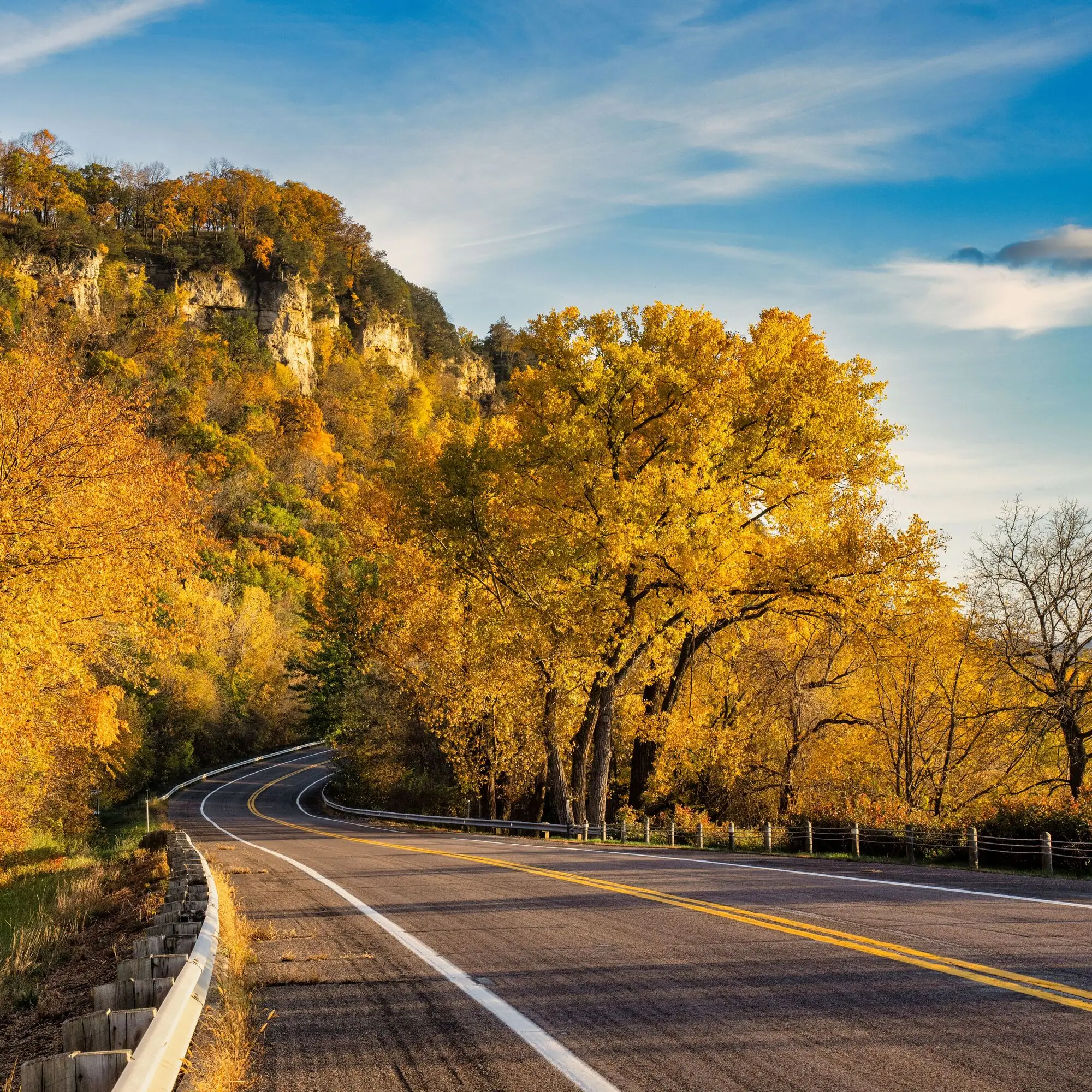 A winding road curves through golden autumn trees; cliffs rise in the background, and the sky is blue—perfect for exploring scenic routes while checking RV rates for your next adventure.