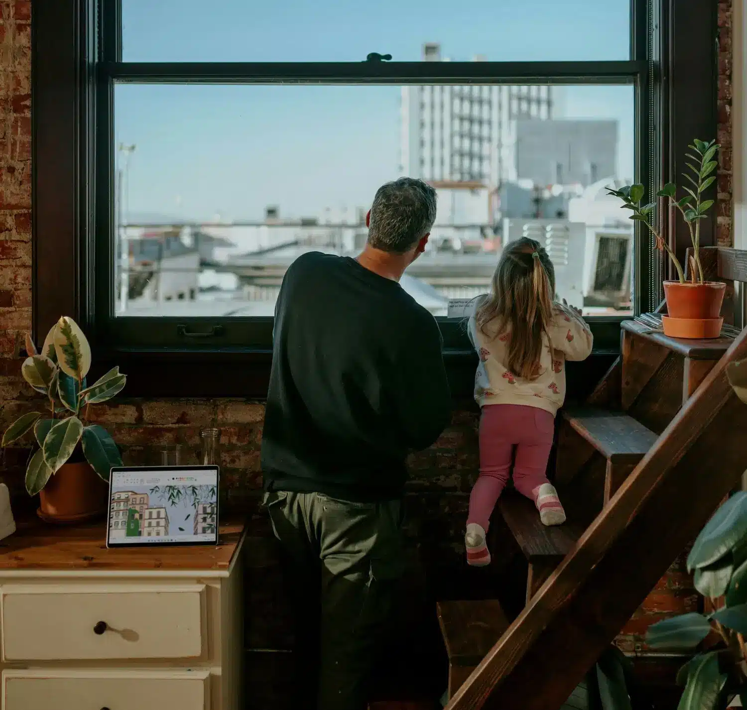 An adult and child look out a window from their loft apartment, where a laptop, drawer, and potted plants hint at dreams—perhaps even plans for credit union savings accounts.