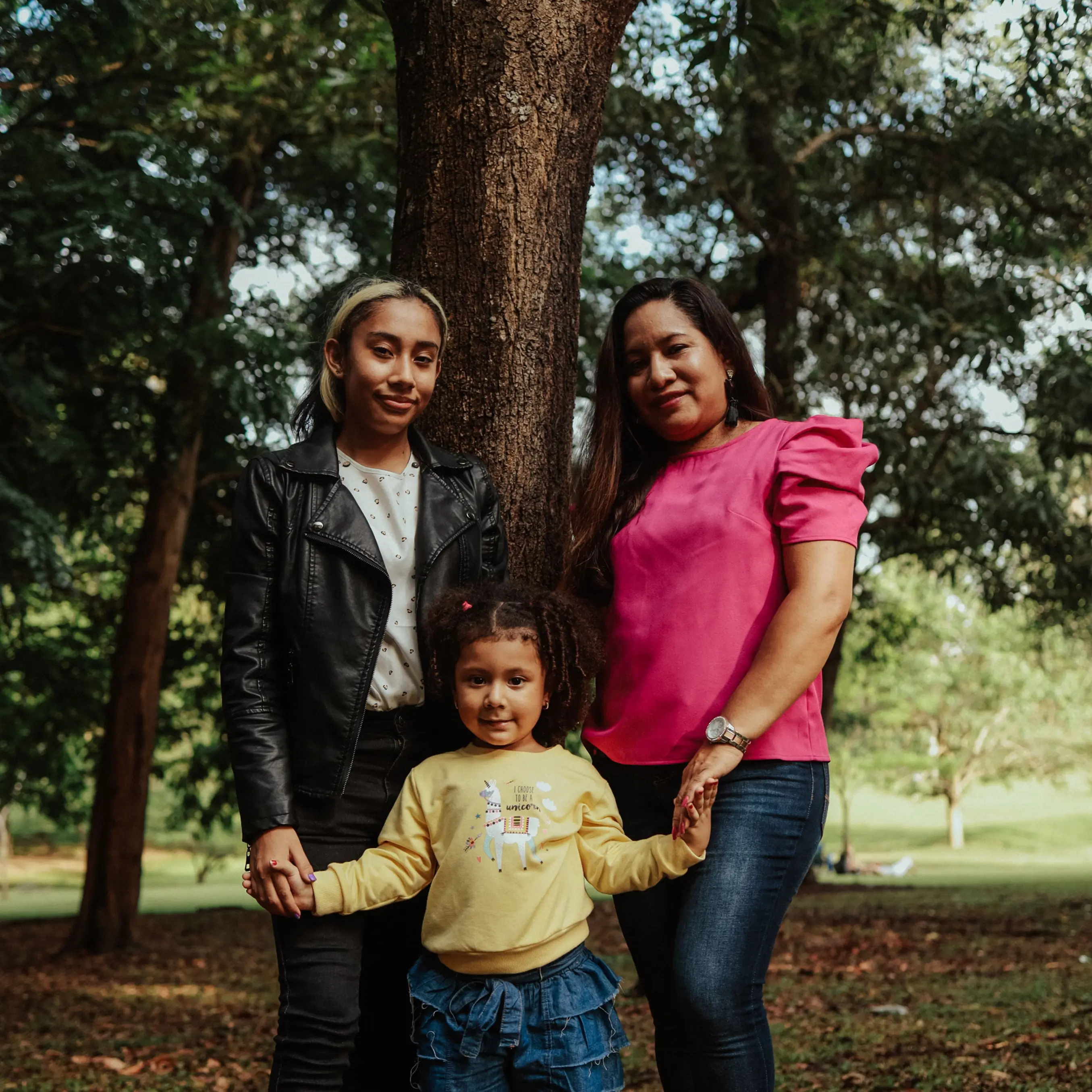 Three people stand together in a park, holding hands. They smile by a large tree, showing the unity often supported by a community credit union.