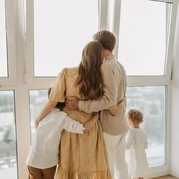 A family of four, seen from behind at large windows of their community credit union, shares a hug as the youngest child gazes outside.