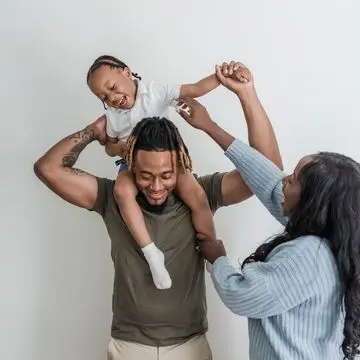 A smiling child sits on an adult’s shoulders while another adult—wearing a community credit union t-shirt—holds the child’s hands. All three appear happy and playful against a plain background.
