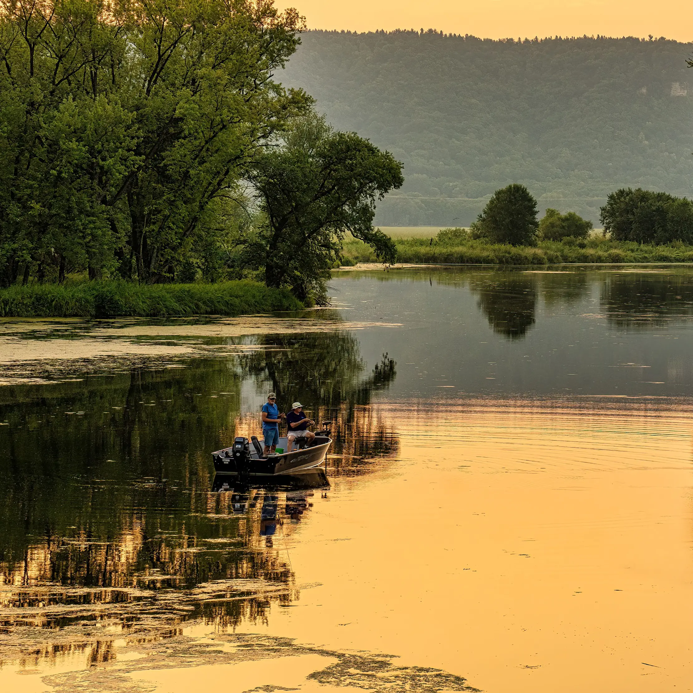 Two people fish from a small boat on a calm, reflective river at sunset, discussing rv rates as they’re surrounded by lush trees and distant hills.