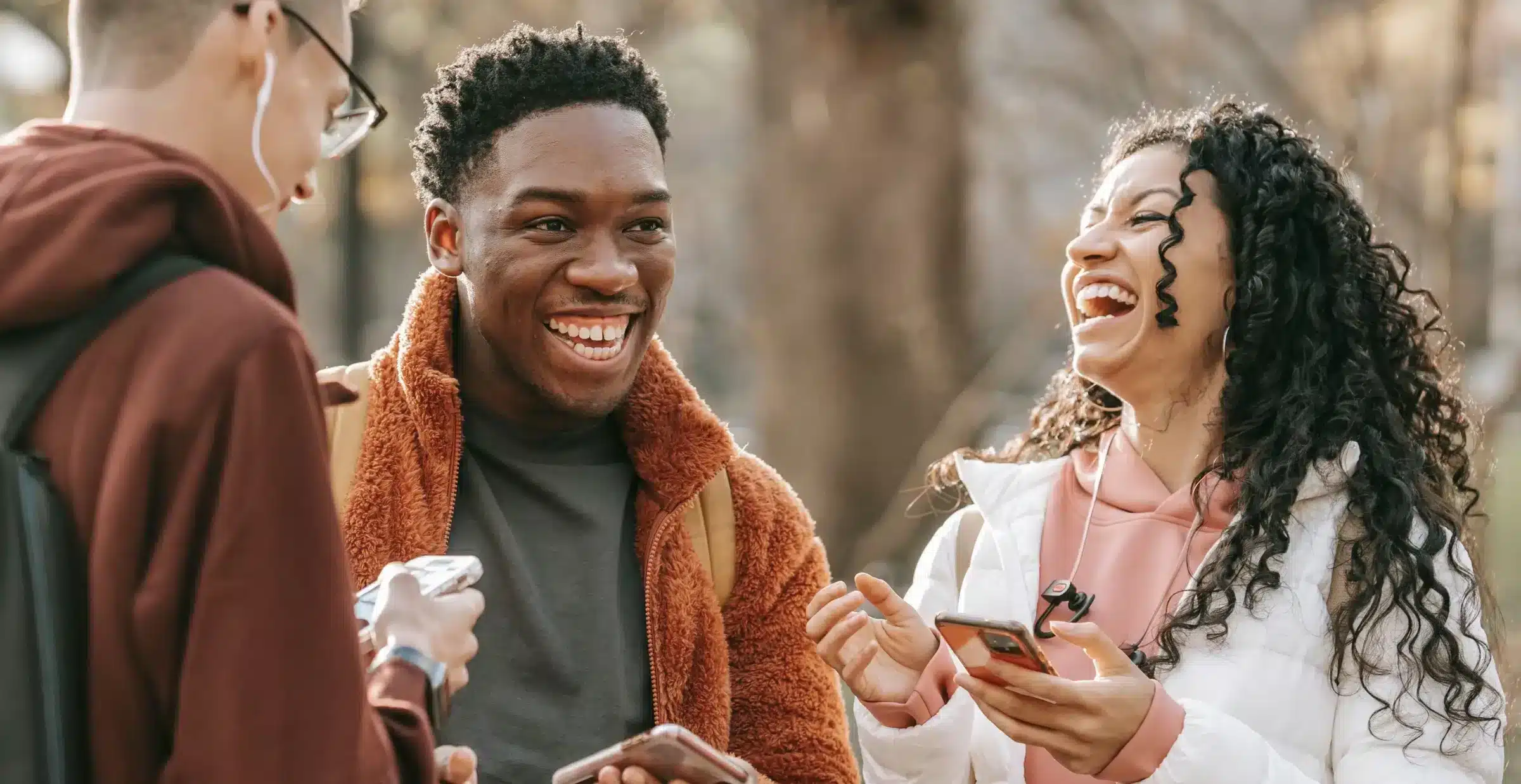 Three young people outdoors, smiling and laughing together while discussing how to join a credit union and enjoying a lively conversation with their phones.