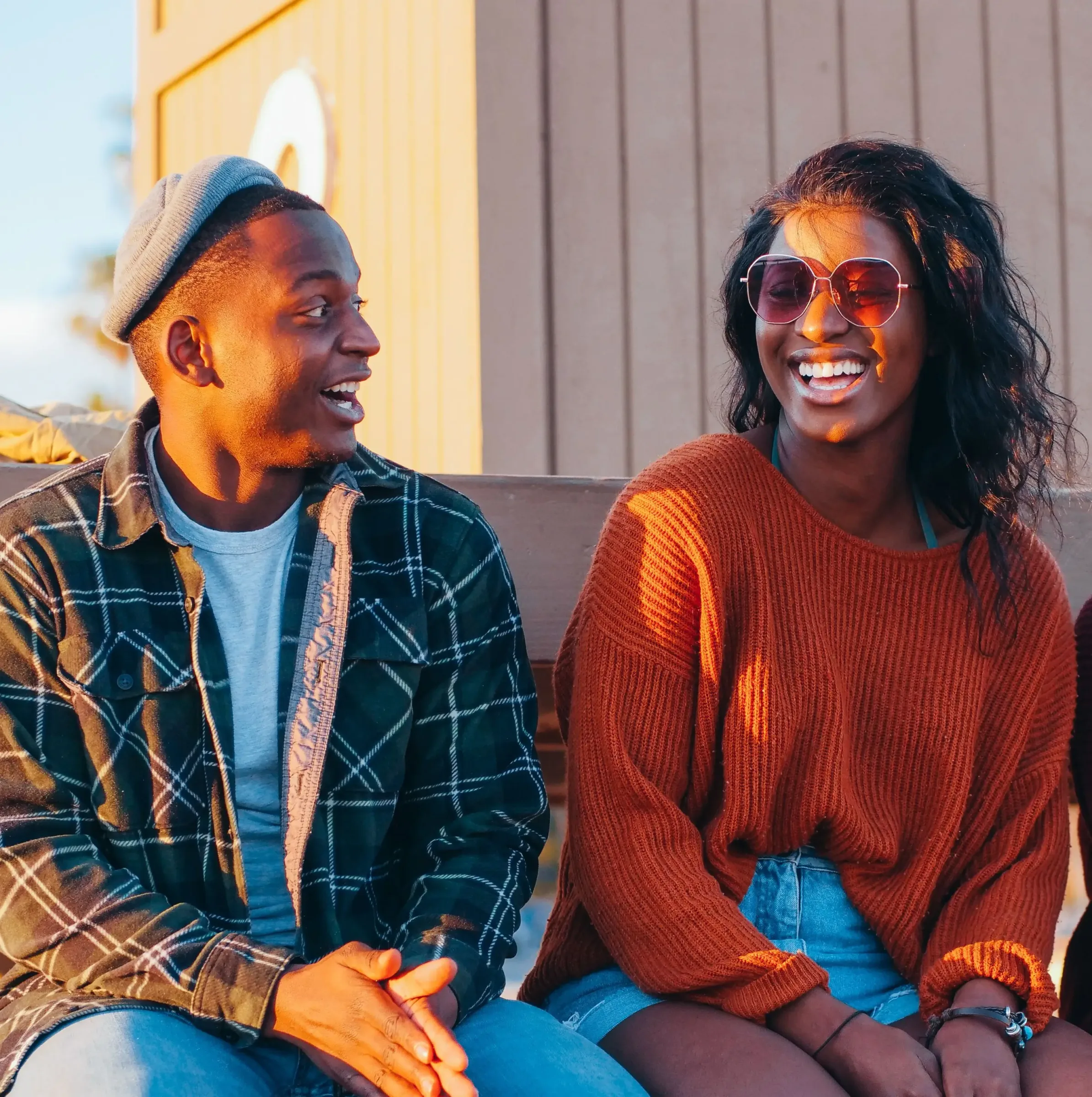 Two people sit outdoors on a bench, smiling and talking about Linden Credit Union. One wears a plaid shirt and hat, the other sports sunglasses, an orange sweater, and denim shorts.