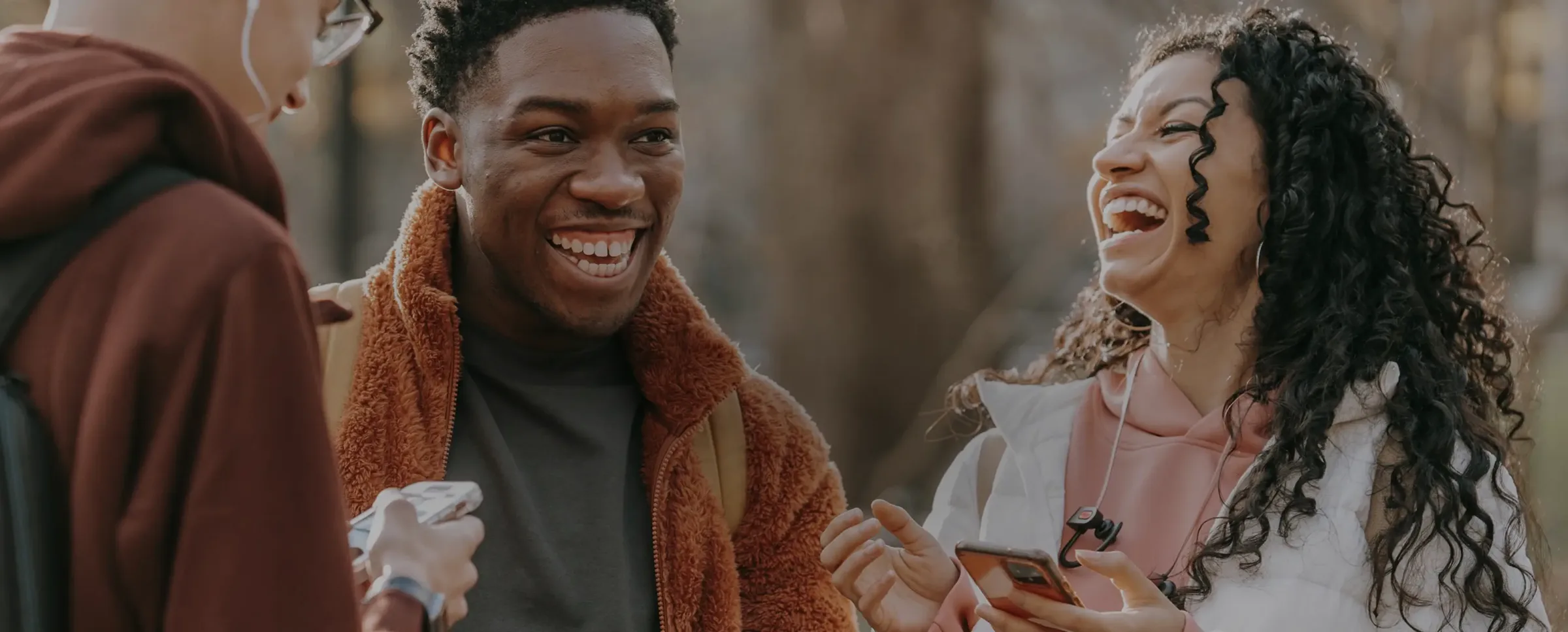 Three young adults laugh together outdoors, chatting and sharing tips on how to join a credit union while holding their phones.