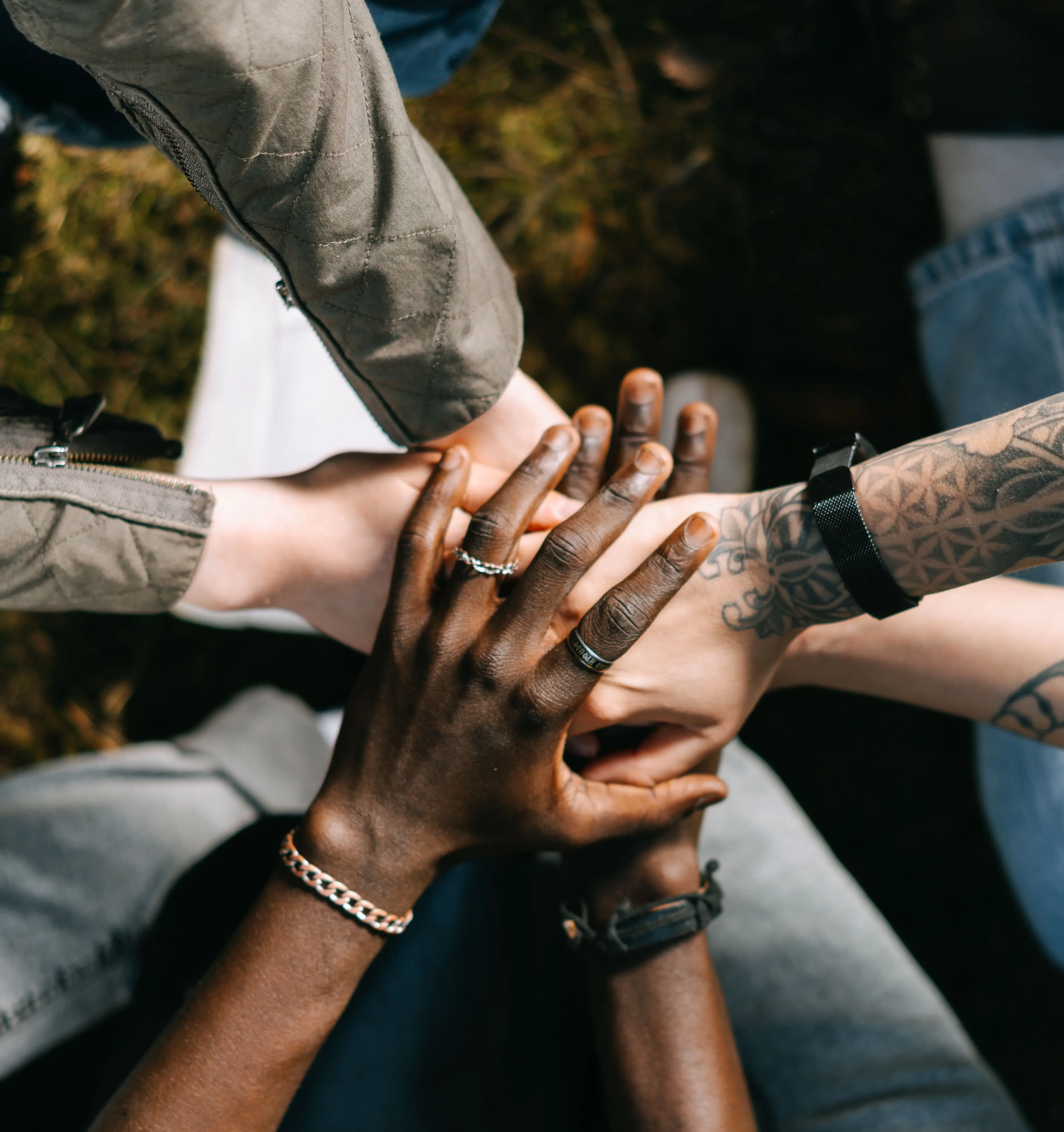 Five hands of diverse skin tones and tattoos are layered on top as they celebrate unity and teamwork, coming together for the saver’s sweepstakes outdoors on grass.