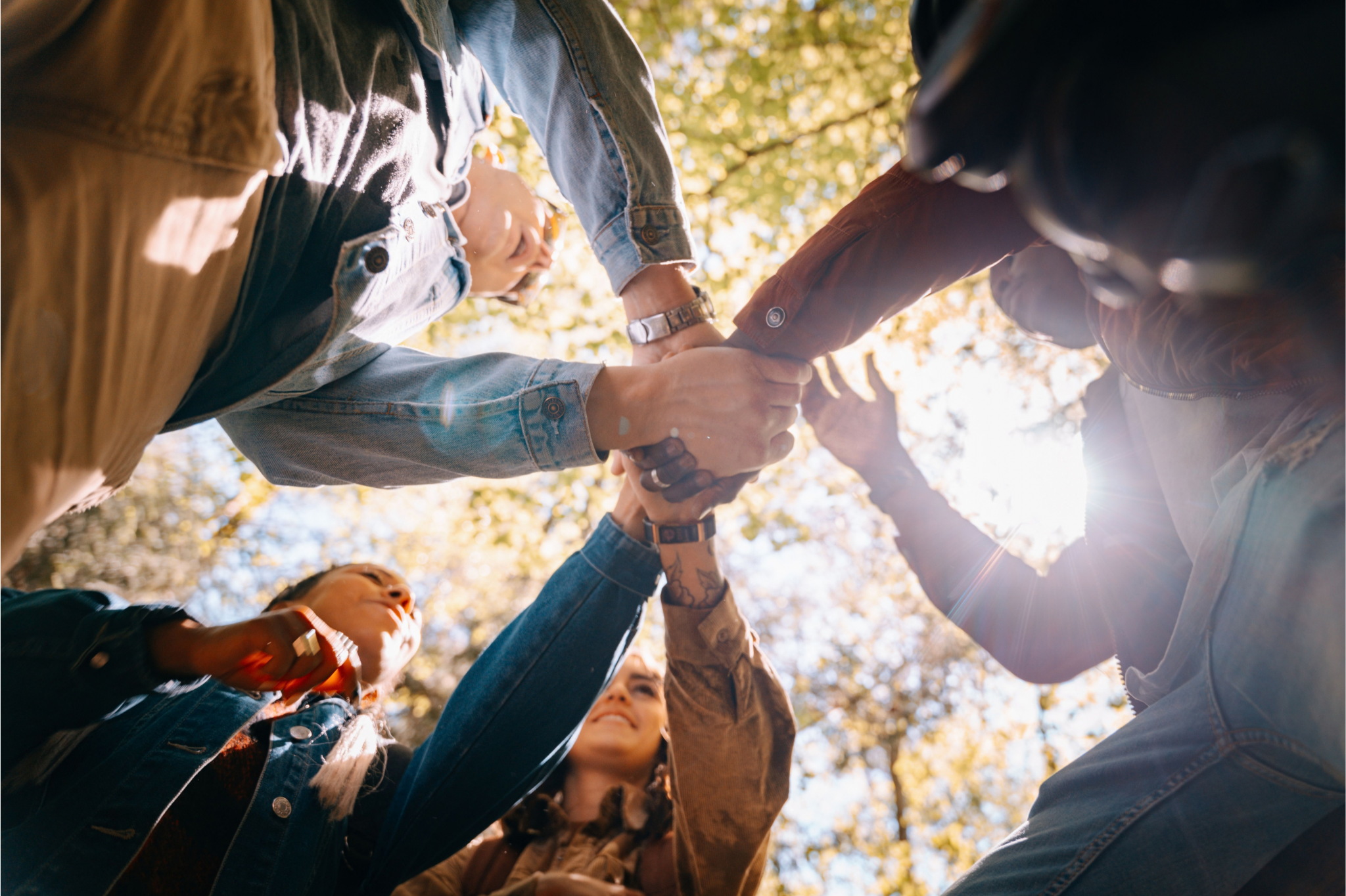 Four people outdoors stand in a circle, reaching hands to the center, showing unity and teamwork. Sunlight filters through trees above them.