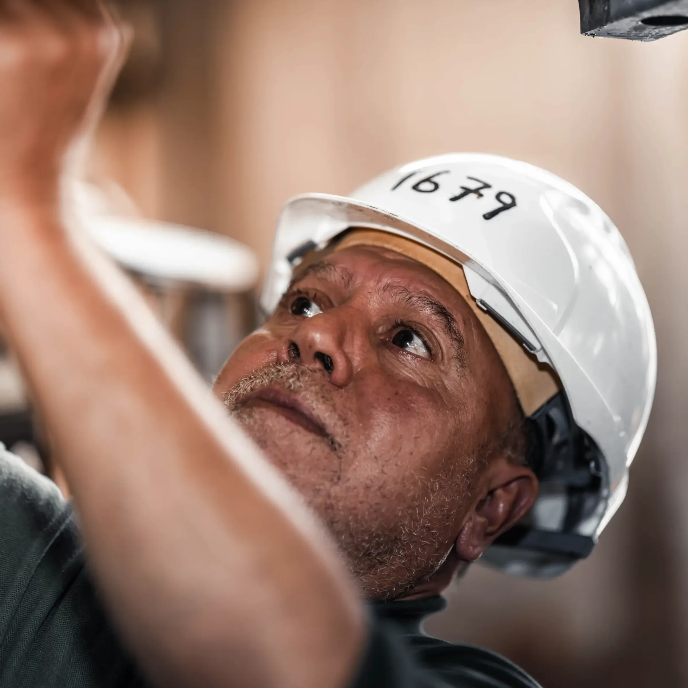 A man wearing a white hard hat labeled 1679 looks up while working, appearing focused—perhaps considering construction loans. The background is softly blurred.