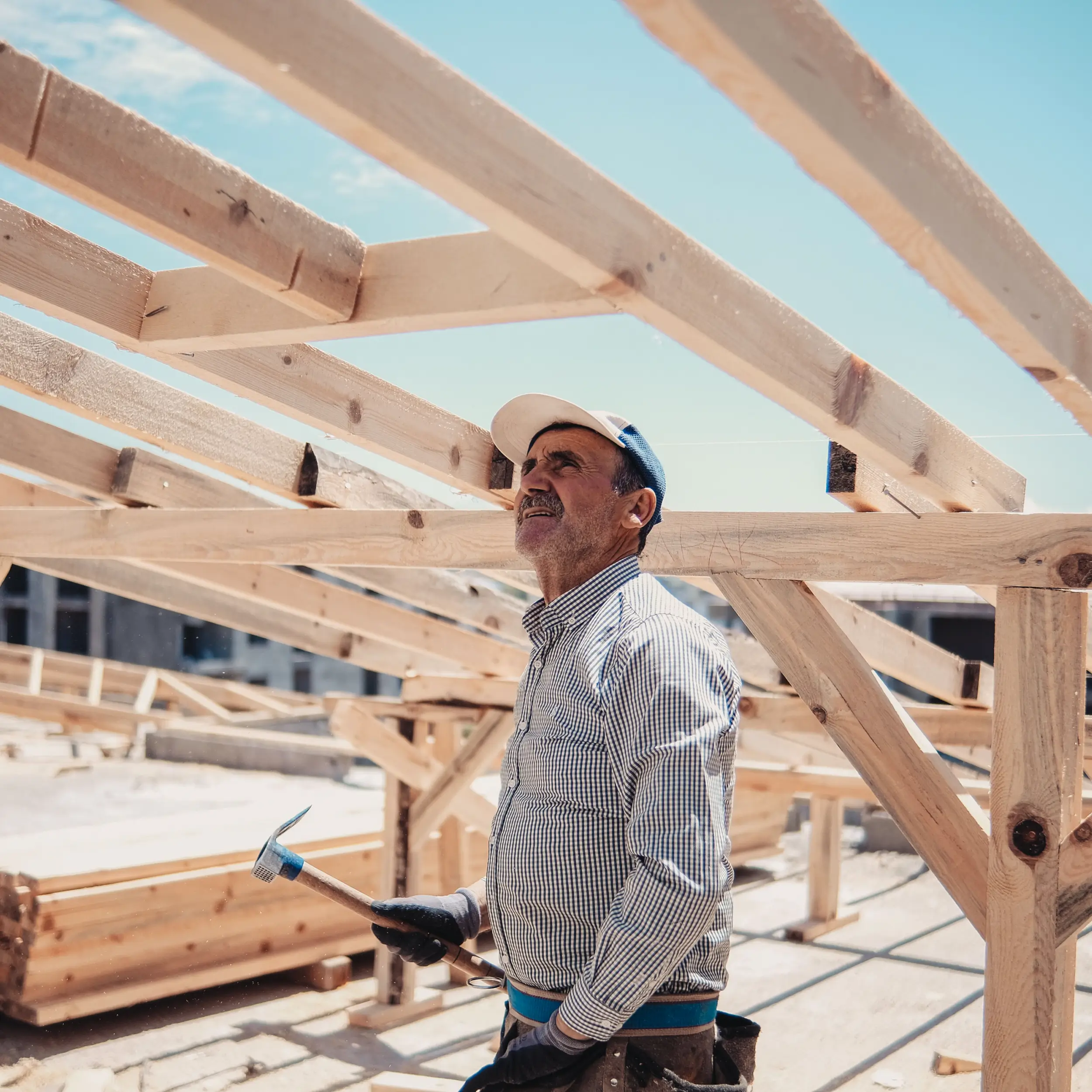 A man wearing a cap and gloves holds a hammer, eyeing wooden roof beams on a construction site under a clear sky—perhaps considering options for construction loans.