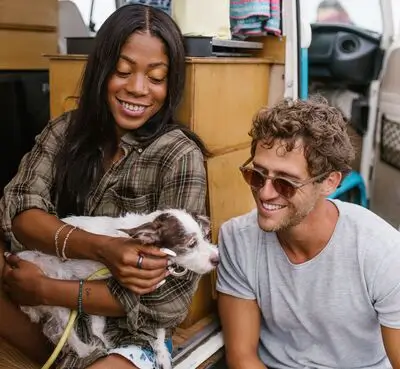 Two people sitting in a van, smiling. One is holding a small white and brown dog on their lap. The scene appears relaxed and happy.