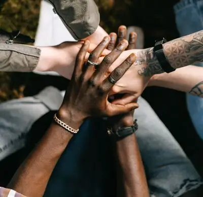 A group of diverse hands stacked together in unity, showing various skin tones, tattoos, and jewelry.