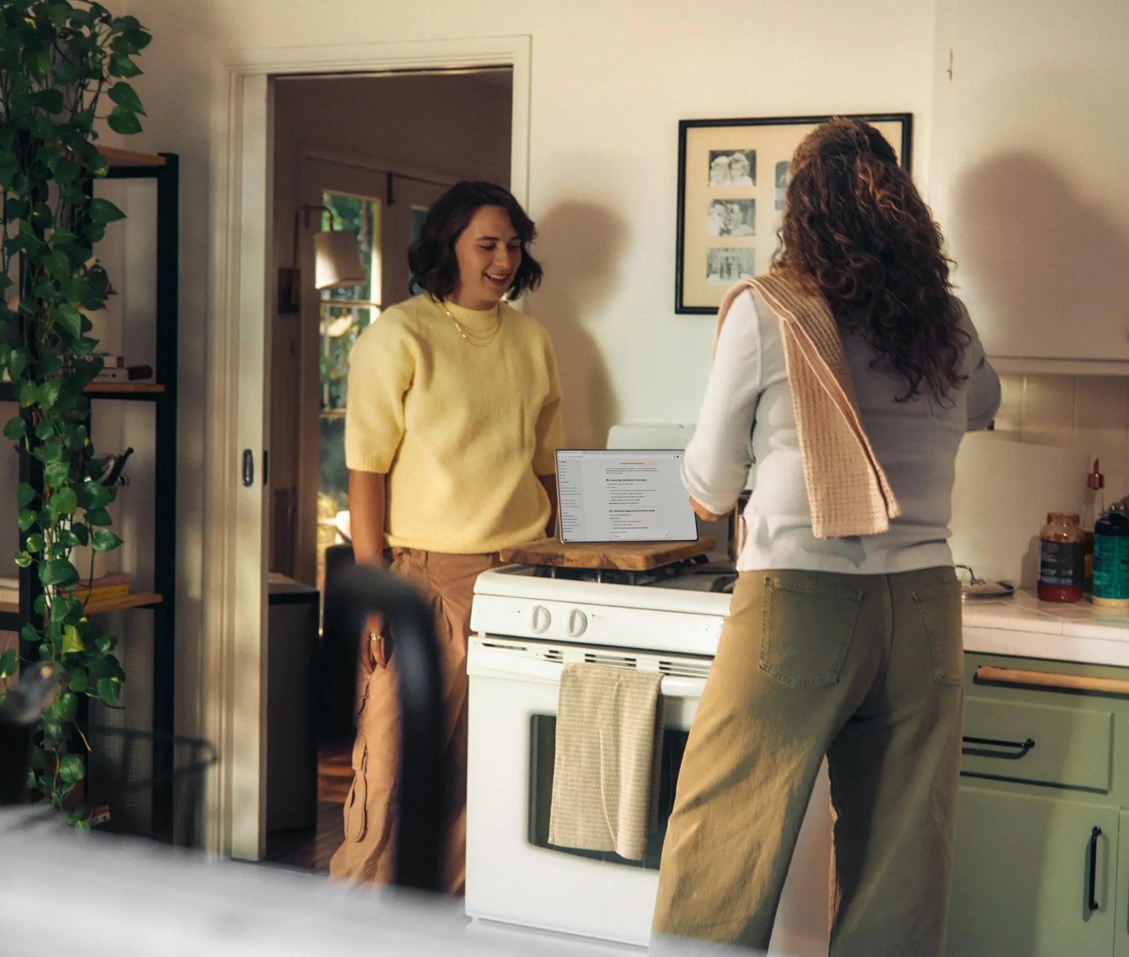 Two people are in a kitchen; one cooks at the stove while the other smiles nearby. Plants, photos, and talk of first time homebuyer loans fill their cozy, newly decorated space.