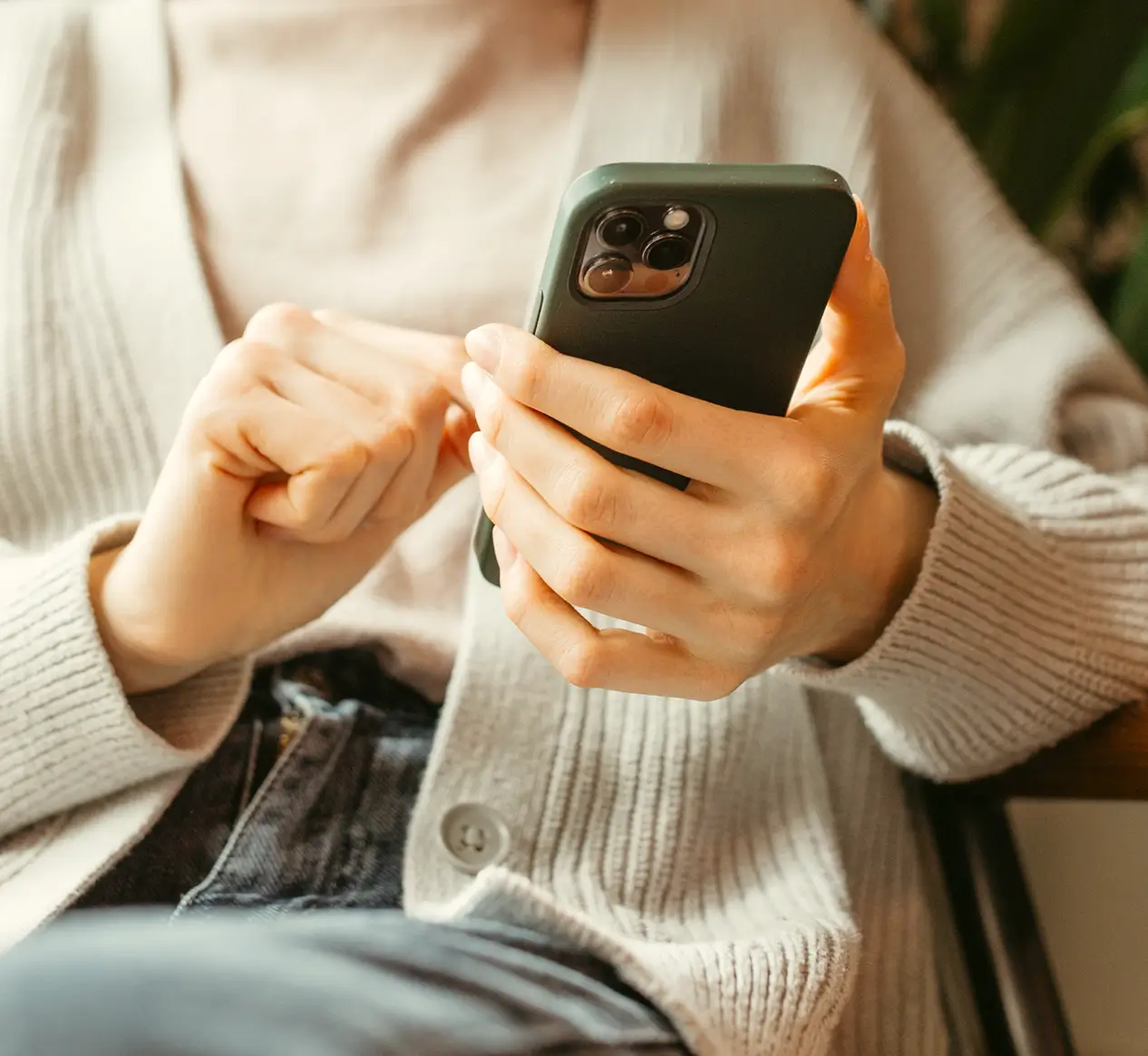 A person in a light sweater and jeans holds a smartphone, researching how to join a credit union, sitting indoors with the focus on their hands and phone.