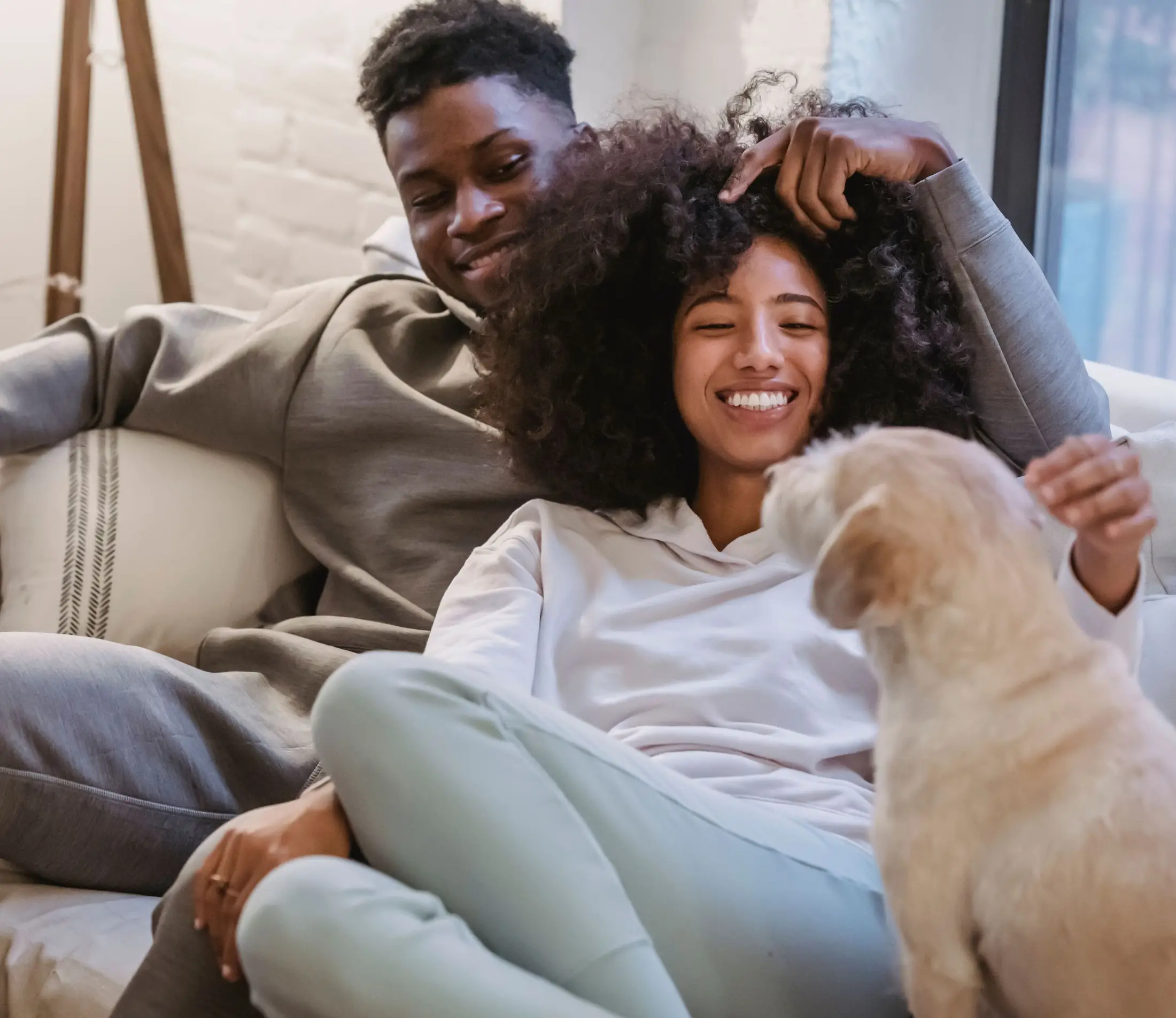 A smiling woman sits beside a man on a couch, petting their small dog—both look happy and relaxed after securing first time homebuyer loans in their cozy living room setting.