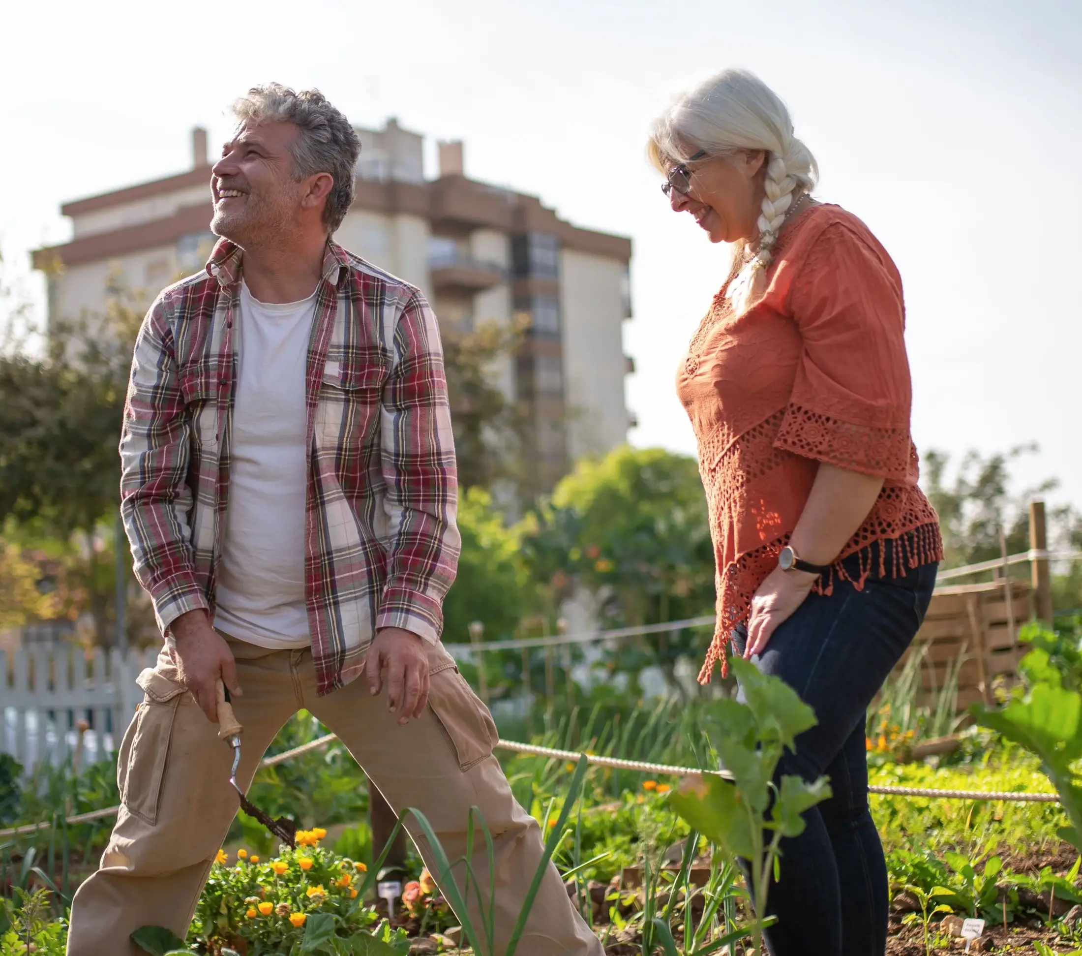 Two older adults smiling and gardening together on a sunny day, enjoying nature’s growth—no credit check loan worries cloud their happiness as buildings and green plants flourish behind them.