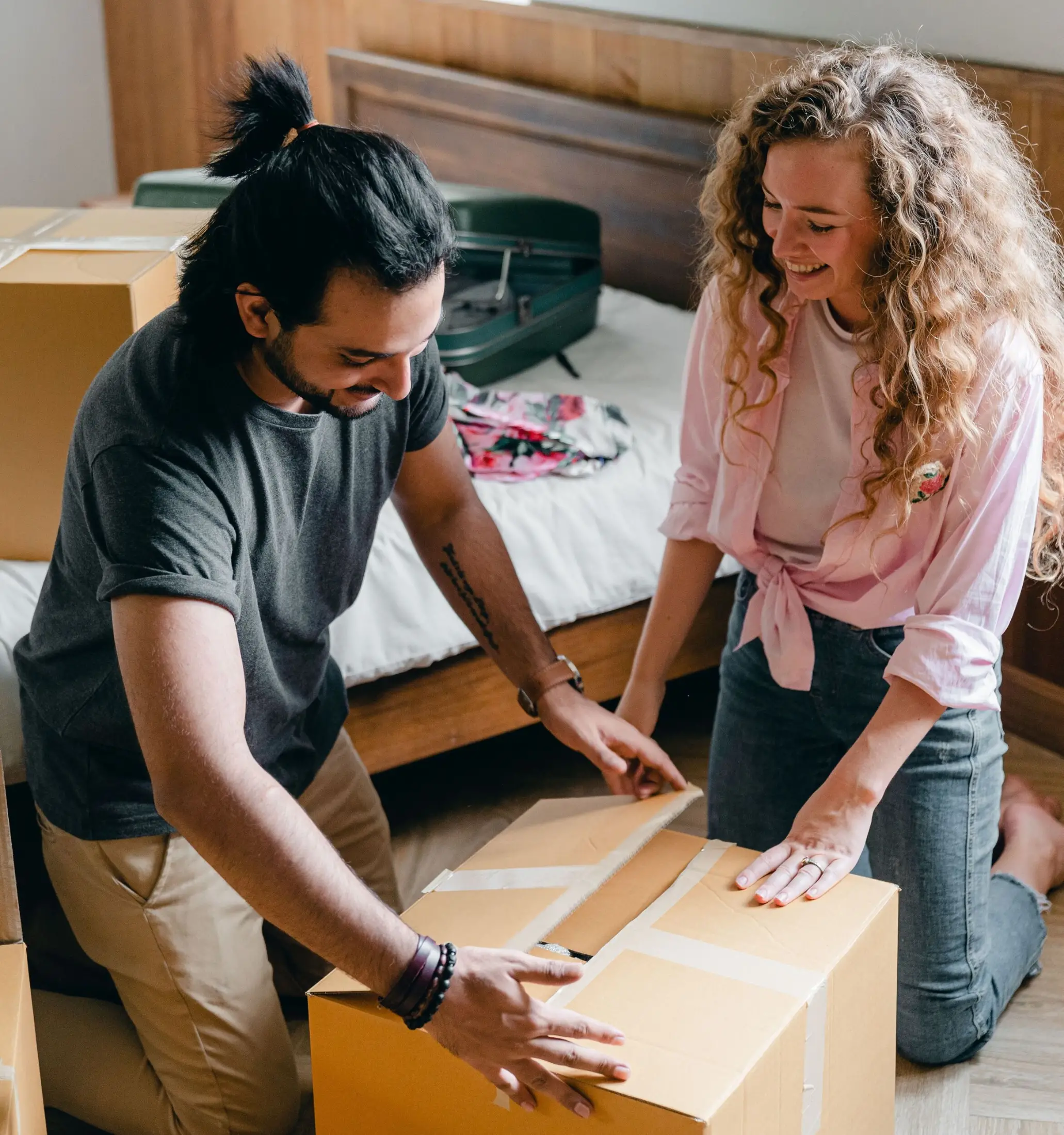 Two people smile while taping a cardboard box in a bedroom, possibly preparing to use their home equity for a move; more boxes and an open suitcase are visible in the background.