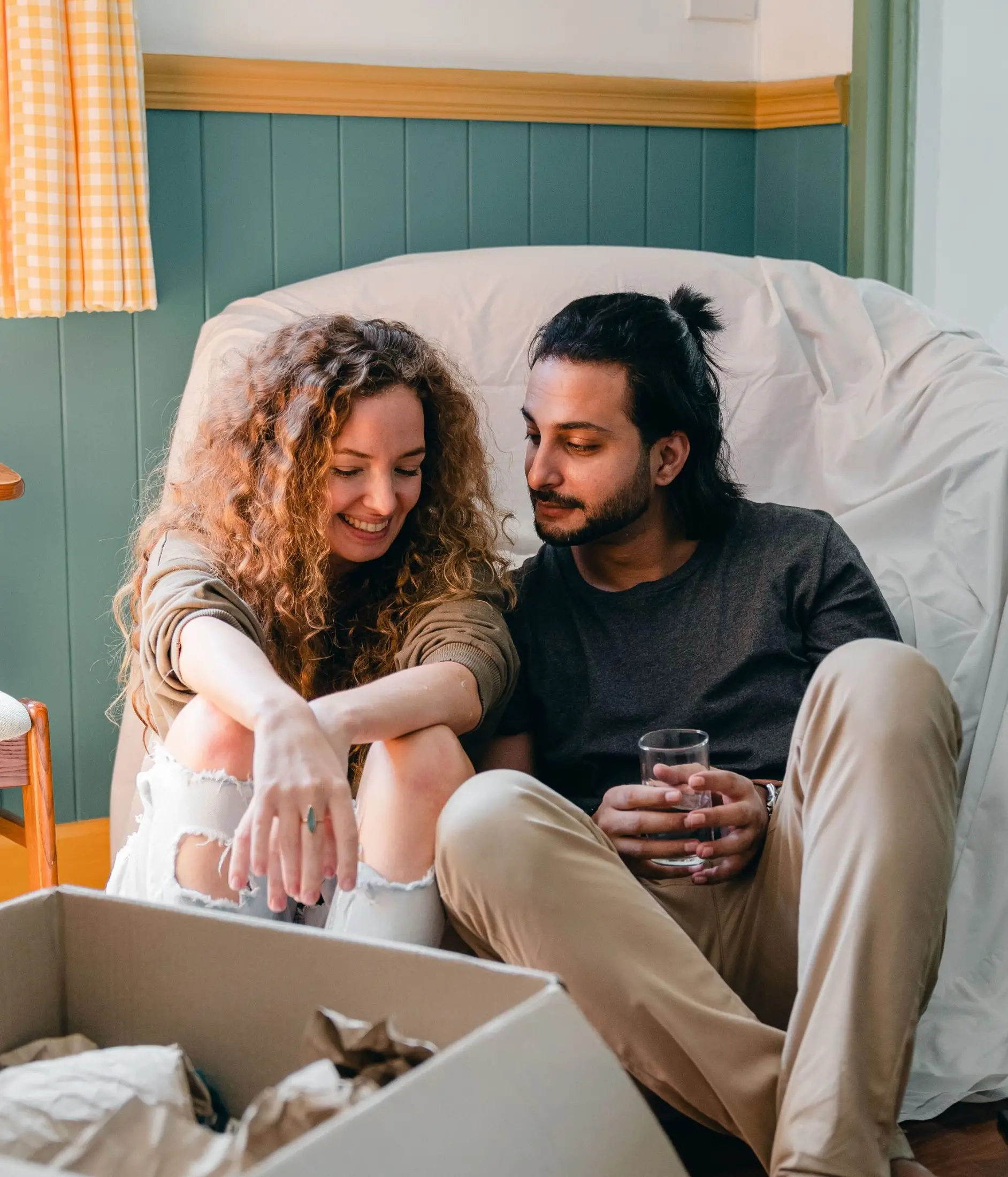 A smiling woman with curly hair and a man holding a glass sit together on the floor by a box, celebrating with first time homebuyer loans in their cozy new room.