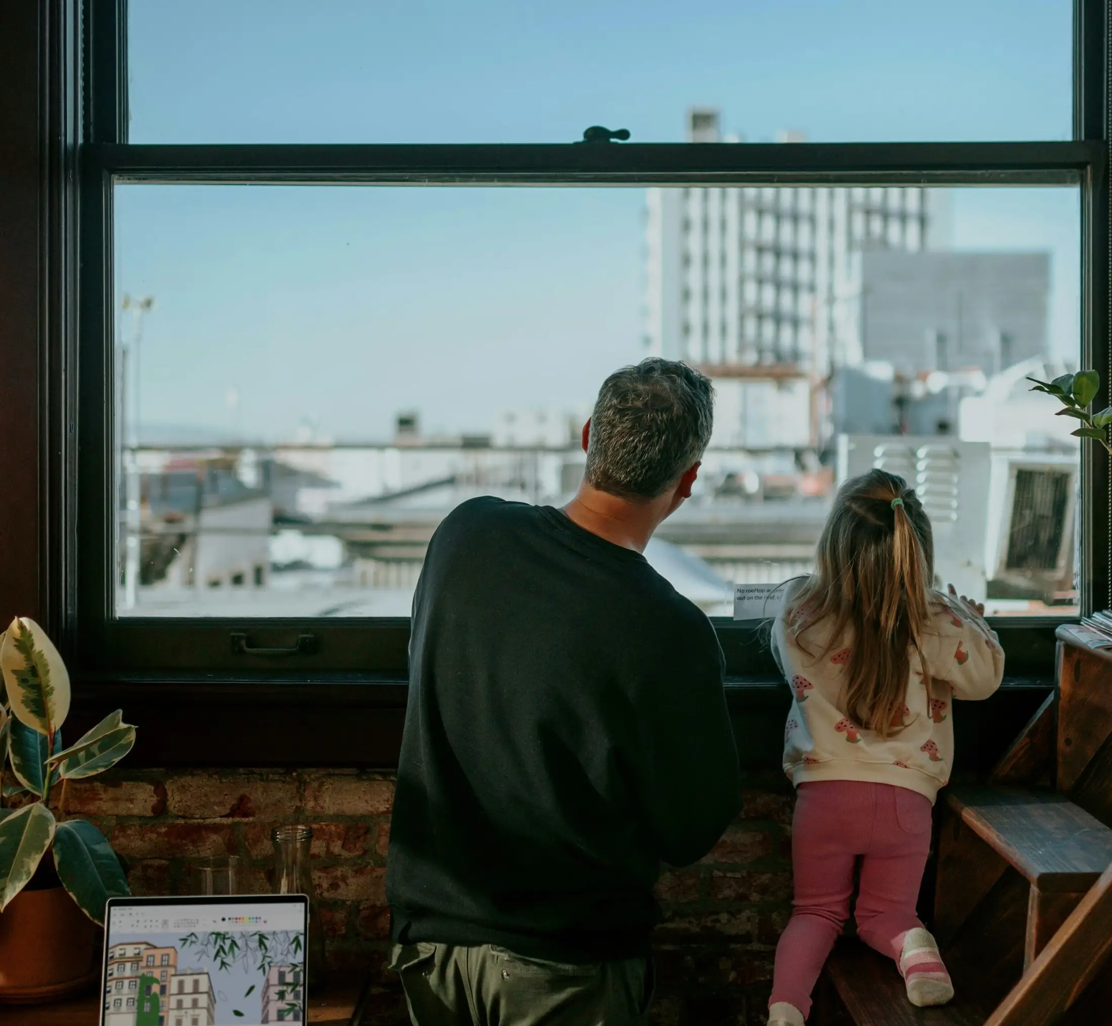 A man and a young girl look out a large window at city buildings, perhaps discussing plans for a share secured loan. A laptop and potted plant sit on the desk nearby.