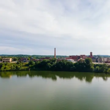 A river reflects trees and a small cityscape, including a tall smokestack and the local community credit union, under a cloudy sky.