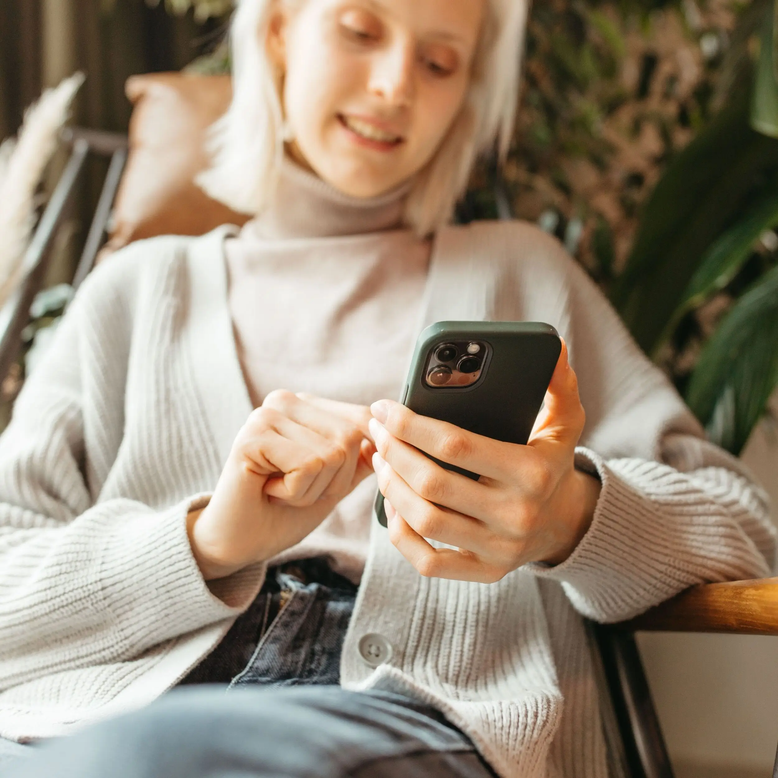 A person in a light sweater and jeans smiles in a chair, using a smartphone to check their credit union checking account, with vibrant plants in the background.