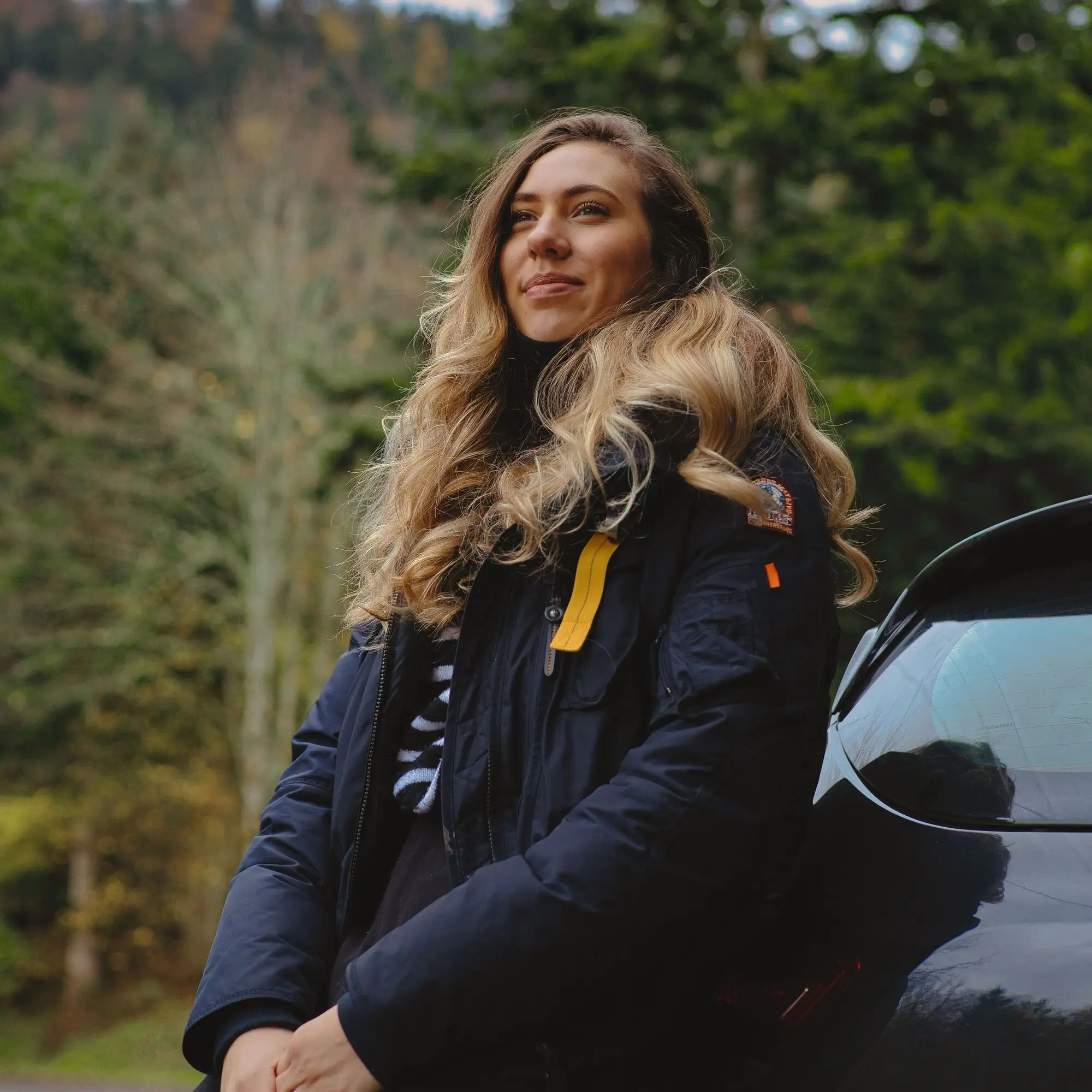 Woman with long wavy hair in a dark jacket stands by a black car, enjoying the view and considering the latest auto loan rates amidst trees and a mountain backdrop.