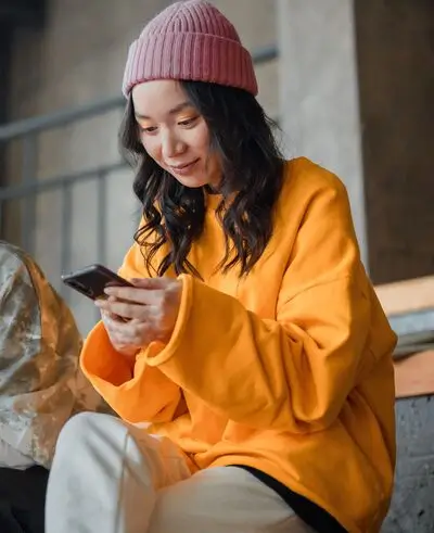 A person wearing a pink beanie and yellow sweater is sitting and smiling while looking at a smartphone.