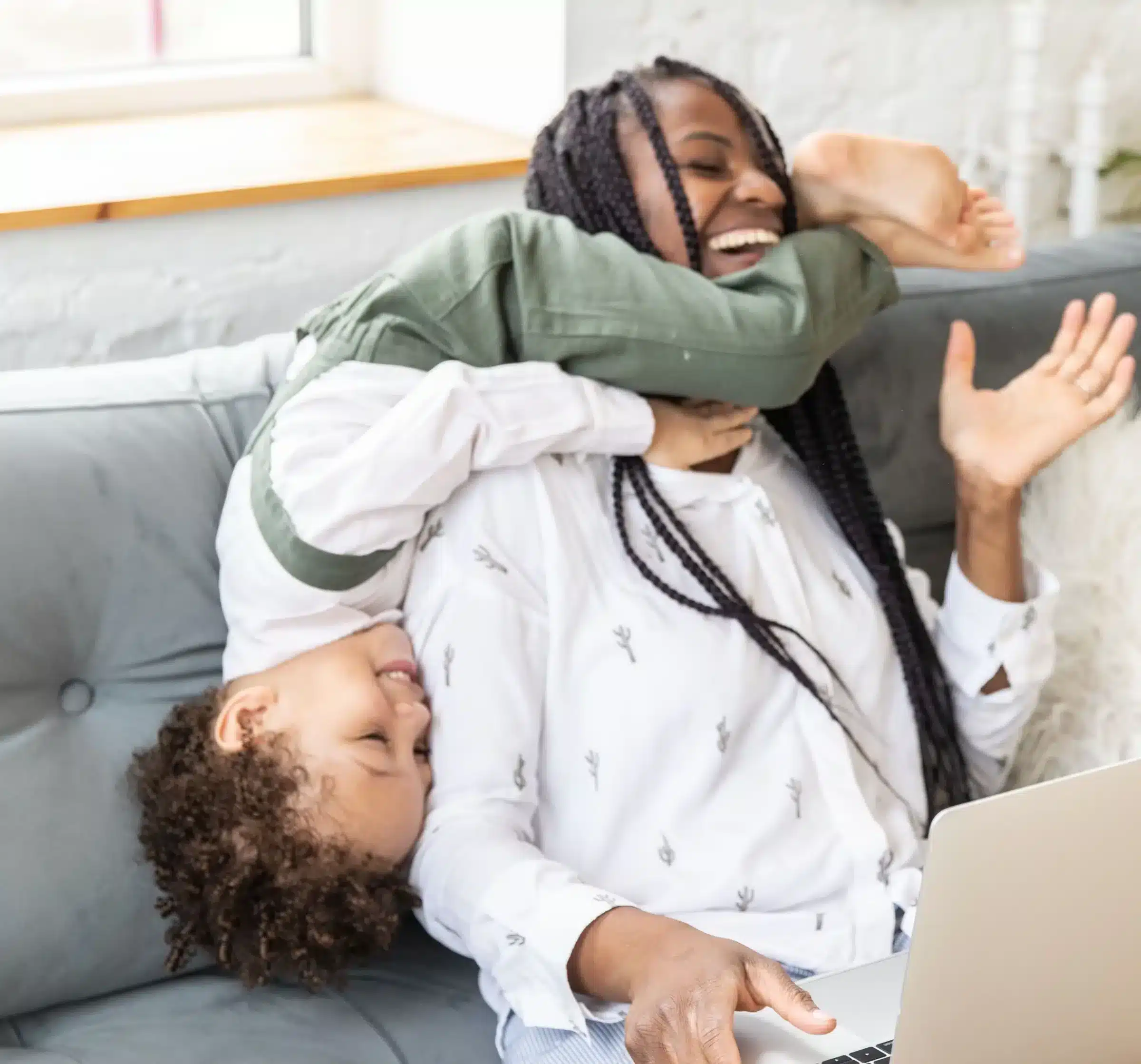 A child playfully hangs upside down on a couch while their mom laughs, working on a laptop and researching how to join a credit union.