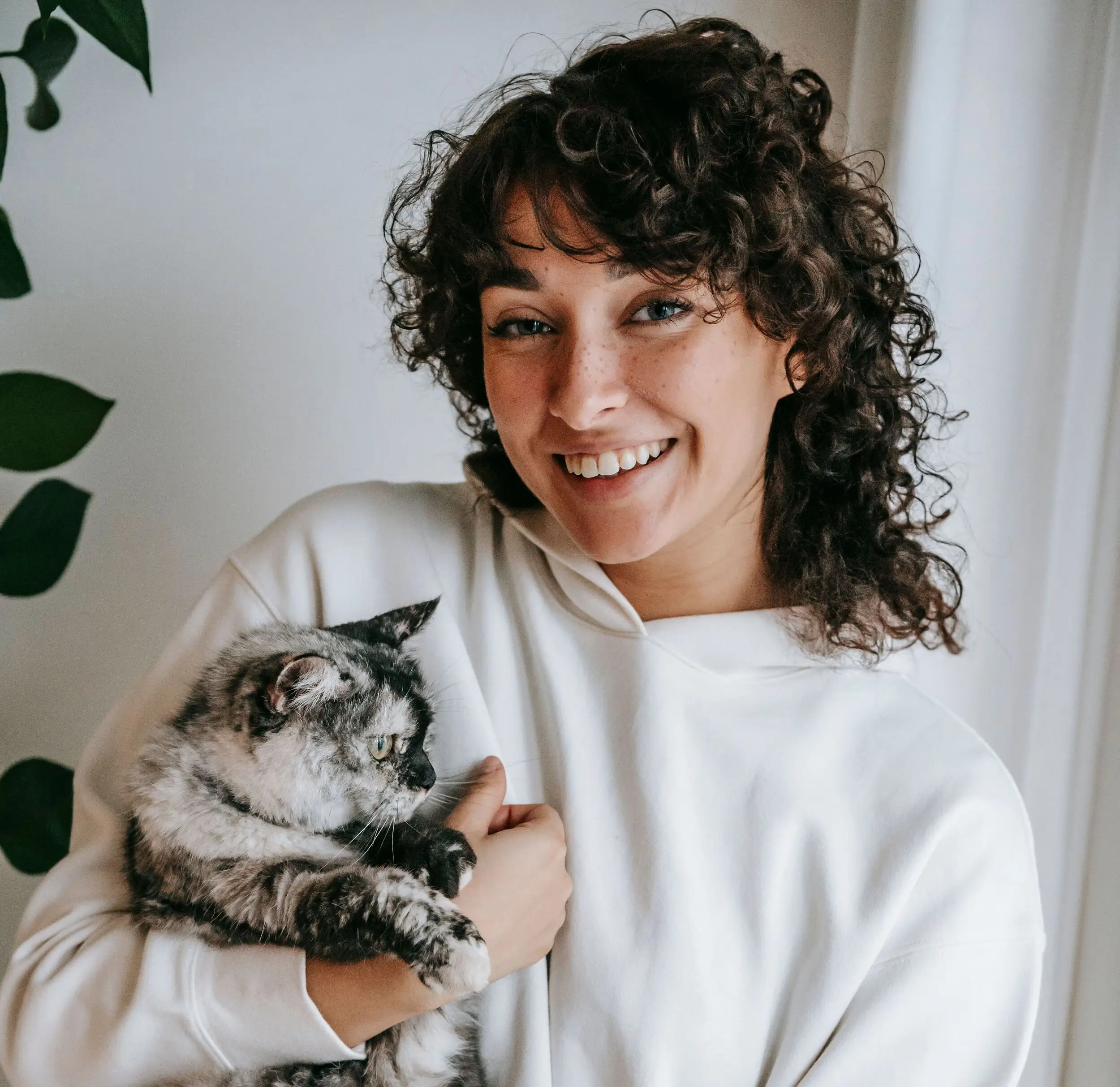 Smiling person in a white hoodie holding a gray and black cat, standing indoors near a leafy plant.