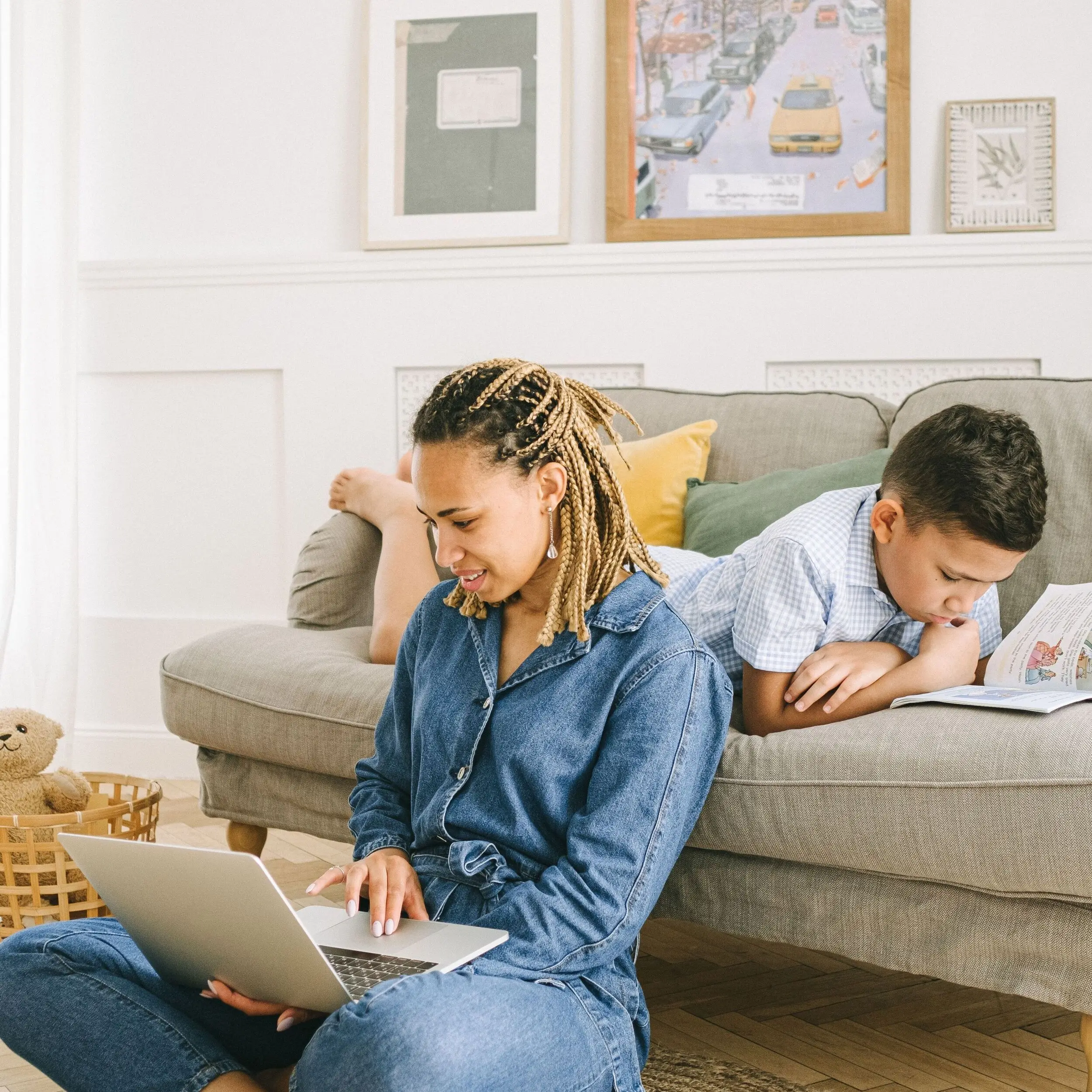 A woman uses a laptop on the floor while a boy reads about youth savings accounts on a sofa in a cozy living room.