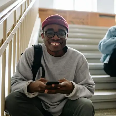 Smiling person wearing glasses, a red beanie, and gray sweater sits on stairs holding a phone, backpack strap over shoulder. Another person is partially visible on the steps behind.