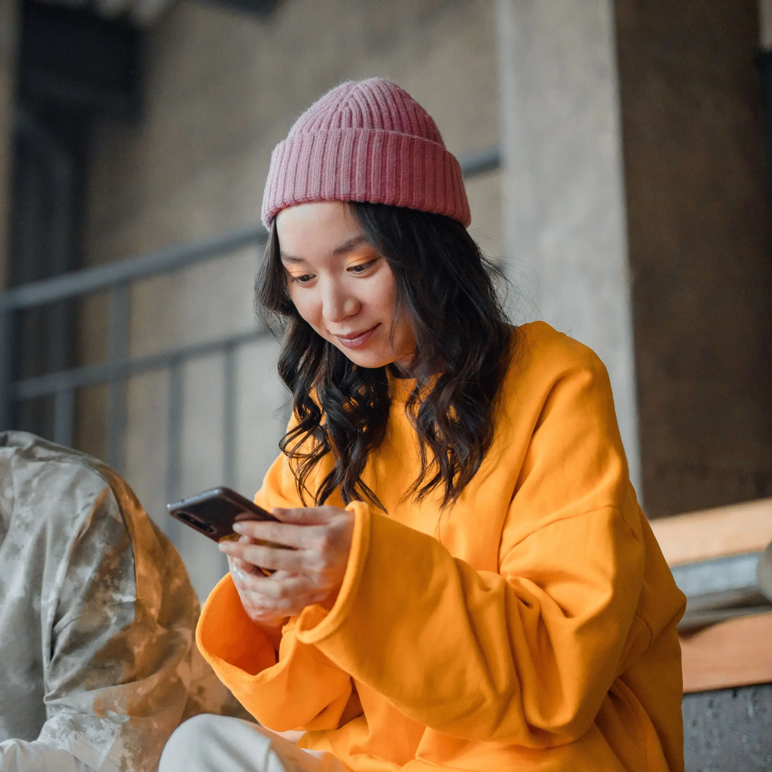 A woman in a pink beanie and yellow sweater smiles indoors, happily checking credit union credit cards offers on her smartphone.